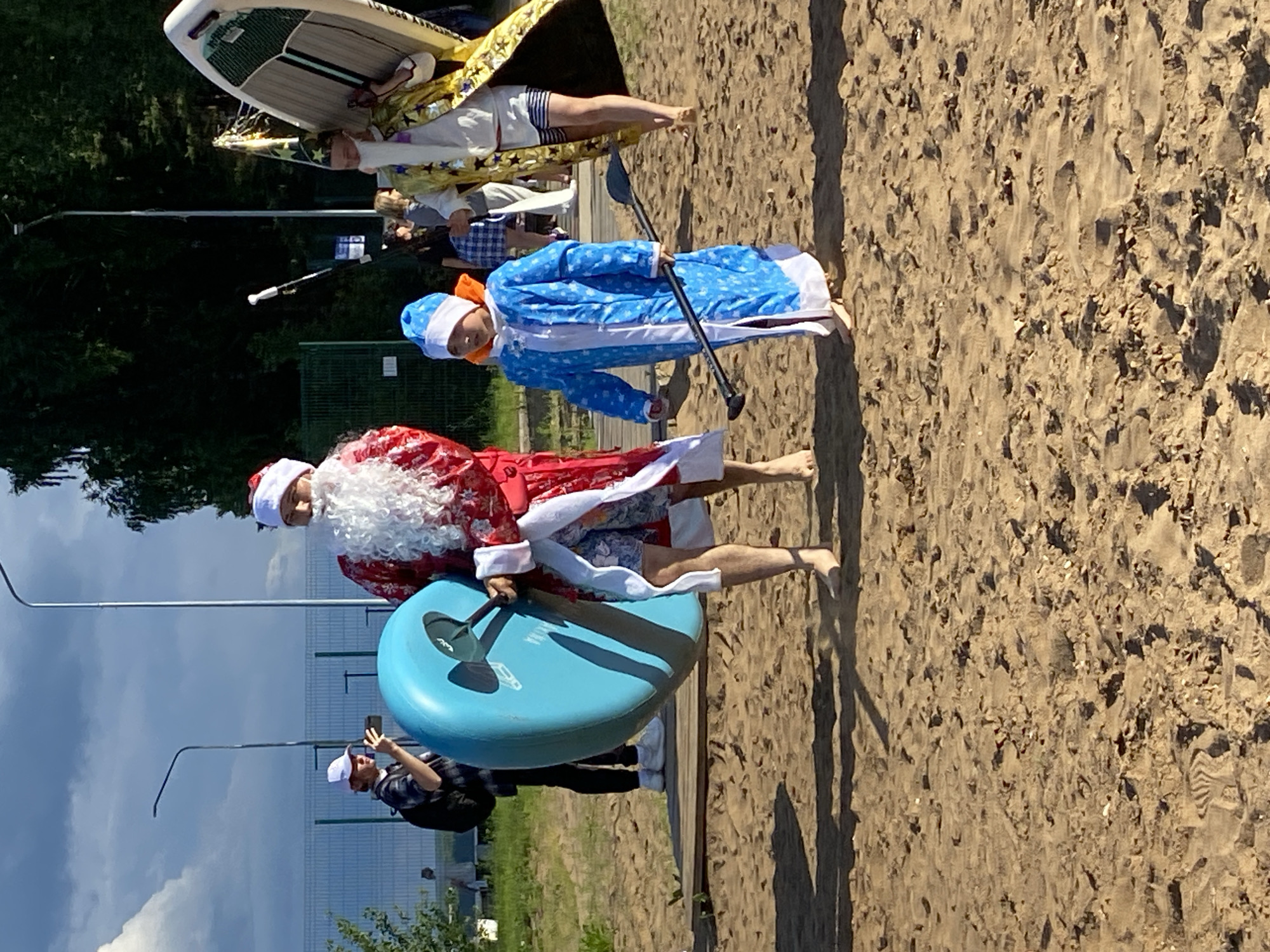 The image depicts a sunny day on a sandy beach, likely at a lake or river, given the presence of paddleboards and a calm body of water in the background. The scene is set in Chaikovsky, Russia. Two individuals are dressed in festive costumes. One person is dressed as Santa Claus, complete with a red suit, white beard, and hat, holding a paddle. They are standing next to a large, bright blue inflatable object, possibly a paddleboard or a giant flotation device. The other person is dressed in a blue and white costume with a hat, also holding a paddle, resembling Snegurochka (the Snow Maiden), a character from Russian folklore who is Santa Claus's granddaughter and helper. Both are barefoot and appear to be participating in some form of paddleboard activity or a themed event on the beach. In the background, other people can be seen, some with paddleboards, and a fence suggests a recreational area. The sky is partly cloudy, indicating a pleasant day. One person in the foreground is holding up a phone, possibly taking a photograph or video. The overall mood appears to be lighthearted and celebratory, fitting for an outdoor event in warm weather.