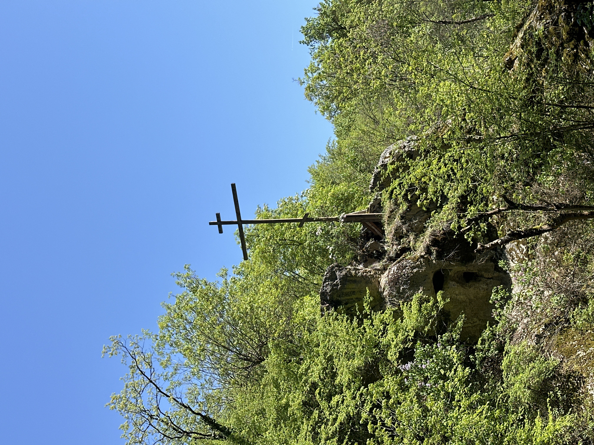 A landscape-oriented photograph shows a wooden cross mounted on a rock formation with green foliage, set against a clear blue sky. The scene is located in the city of Lilyache, Bulgaria. The cross is positioned off-center to the left, appearing dark against the sky. The rock formation is covered in green plants, and surrounded by trees with green leaves. The sky is a uniform blue, devoid of clouds. The photograph appears to be taken from a low angle.