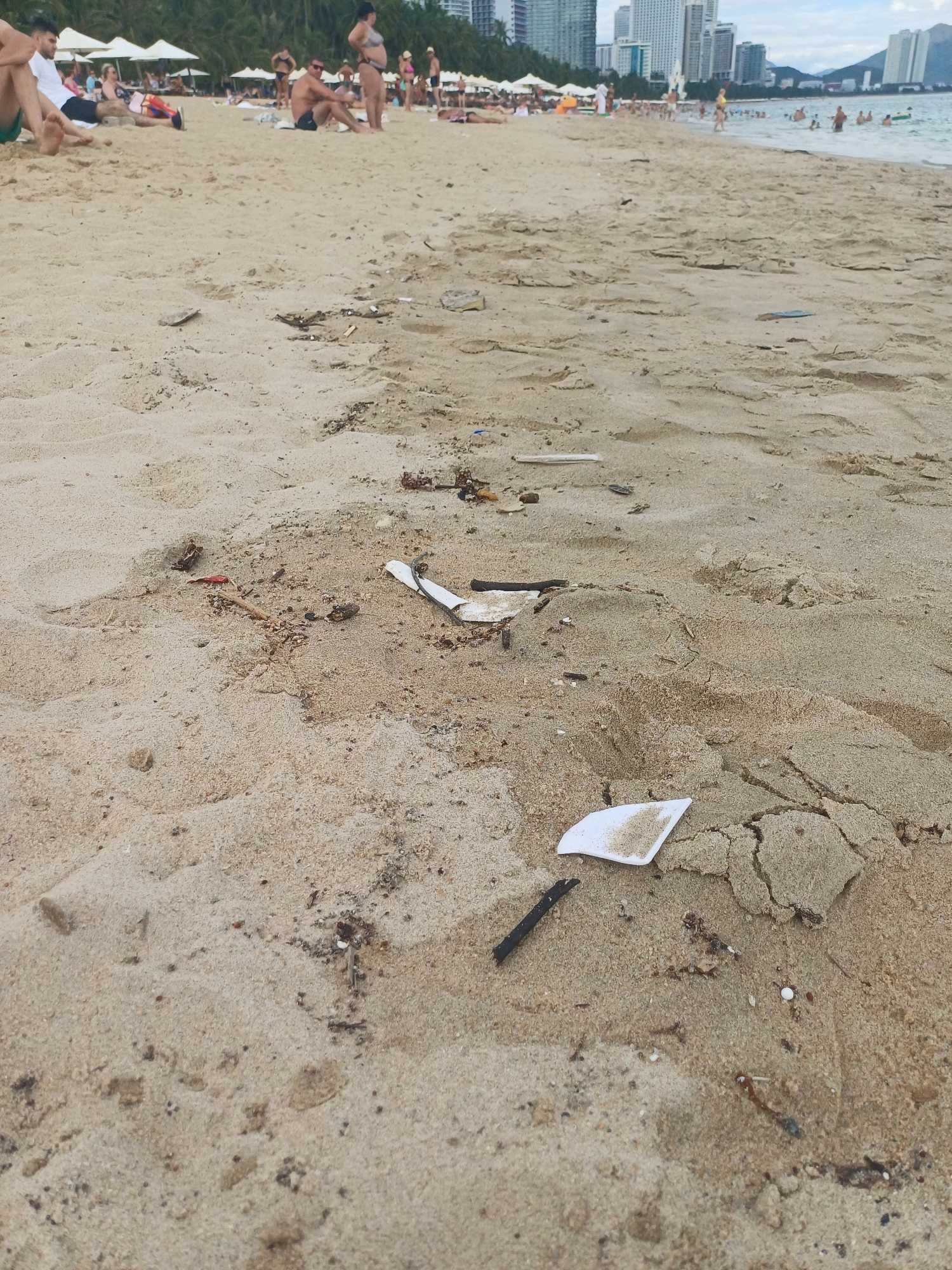 The image depicts a sandy beach in Nha Trang, Vietnam, with several people in the background enjoying leisure activities. The foreground of the beach is littered with debris, including pieces of white plastic, dark twigs, and other small, unidentifiable objects. A few small, dark brown specks, possibly dirt or dried organic matter, are scattered across the sand. In the mid-ground, people are seen sitting and standing on the sand, some under white beach umbrellas. The ocean is visible in the background with people swimming and the coastline is lined with palm trees and modern high-rise buildings, characteristic of Nha Trang's urban coastal landscape. The sky is overcast.