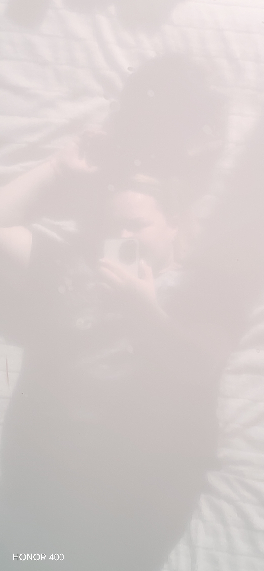 An overexposed and washed-out mirror selfie captures a person indoors, likely in Noviy, Russia. The image is very hazy, with most details obscured by bright white light.
The primary subject is a person, likely an adult, appearing to be female based on the faint facial outline and hair. They are taking the photo with a light-colored smartphone or small camera, held up to capture their reflection. The person has what appears to be dark, somewhat wavy hair. They are wearing a dark-colored top or dress.
The setting appears to be an indoor room, possibly a bedroom. The background behind the person's reflection shows a light-colored, textured surface, perhaps a white or cream bedspread or wall covering with subtle patterns or ruffles.
The overall image quality is poor due to severe overexposure, creating a dreamlike or ethereal effect where features are barely discernible. Small, faint circular light artifacts or dust motes are visible in various parts of the reflection. In the bottom left corner, the text "HONOR 400" is visible, indicating the model of the camera used to take the photograph or a watermark from the device.