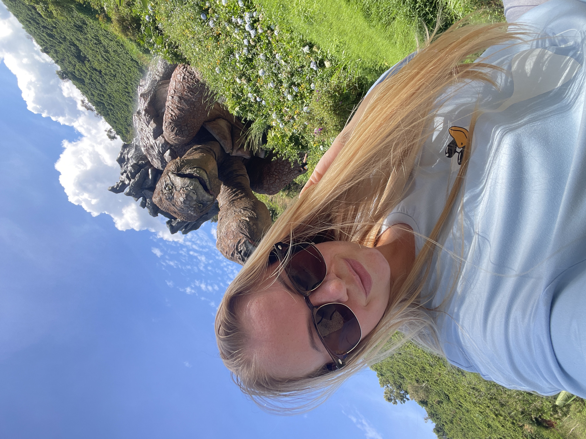 A blonde woman with sunglasses and long, blonde hair is taking a selfie outdoors. She is wearing a light blue t-shirt with a small yellow duck graphic on it. Behind her, a large, realistic sculpture of a tortoise is situated on a grassy hillside dotted with small white flowers. The background consists of a lush green, tree-covered slope under a bright blue sky with scattered white clouds. The scene appears to be a tourist attraction in Thôn 1, Vietnam.