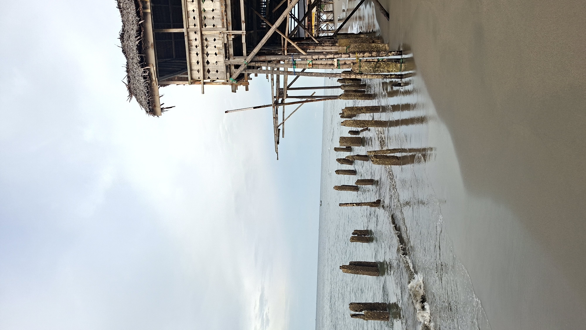 The image captures a view of an abandoned wooden pier extending over a sandy beach and the ocean. The pier is constructed of weathered wood, with a thatched roof visible on the left side. Numerous wooden pilings, encrusted with barnacles and seaweed, jut out of the water along the shore, creating a textured foreground. The sea is calm with gentle waves lapping at the beach. The sky is overcast, suggesting a cloudy or possibly early morning or late afternoon time of day. The overall scene evokes a sense of decay and abandonment, with the natural elements beginning to reclaim the man-made structure. There is no visible text in the image. The location context provided is Lhokseumawe, Indonesia, which aligns with the tropical beach setting and weathered wooden structures often found in coastal areas of Southeast Asia.