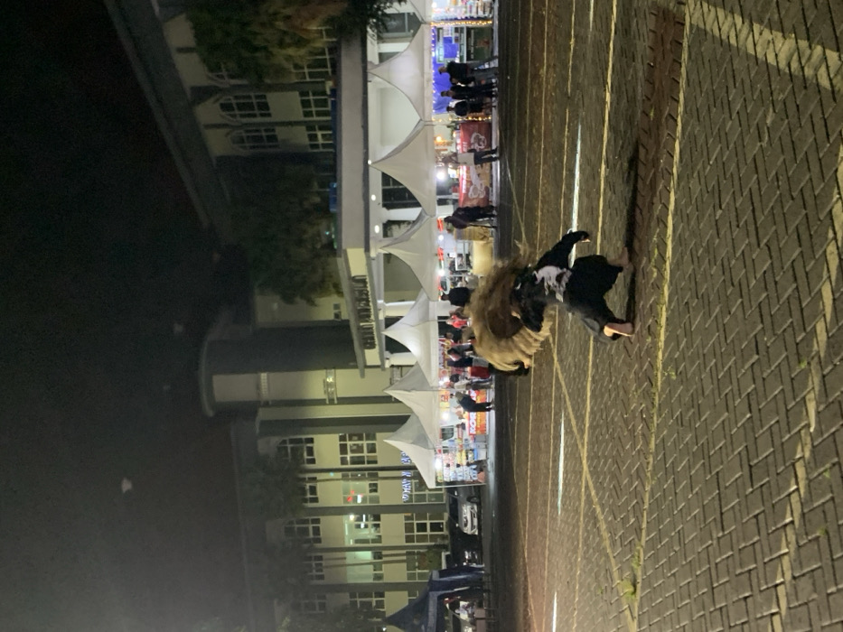 A man is performing a traditional dance in a paved parking lot in front of a large building in the city of Kecamatan Batu, Indonesia, at the coordinates -7.867 latitude and 112.514 longitude. The man wears a black shirt and pants, and a large, brown, fluffy headpiece. He is crouched down, with his arms extended, as if he is about to leap. The man's dance is performed in front of a building with white tent-like canopies set up in front of it. The canopies appear to be part of a market or festival. The building is two stories high and has many windows. The parking lot is empty except for the performer and a few people in the distance. The scene is illuminated by overhead lights. There are white lines painted on the parking lot. The building is in the background and the parking lot is in the foreground. The man is the only person in the foreground of the image. The night sky is visible in the background, and there are streetlights on the building.