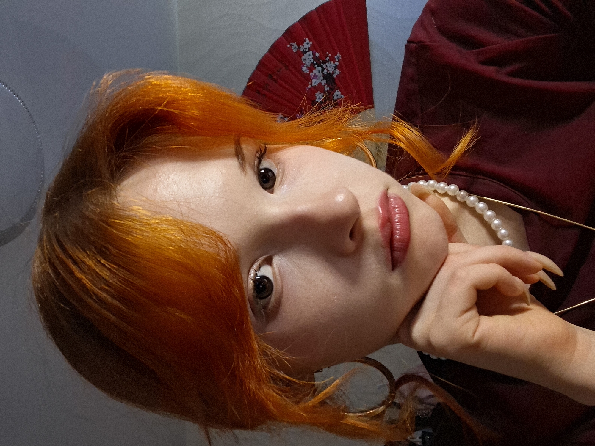 A portrait of a young woman with bright orange hair, pale skin, and brown eyes. She is wearing a string of pearls and a dark red top. Her left hand is positioned under her chin. A red fan with a white cherry blossom design is visible behind her head. The background appears to be a wall of a light grey color, and the photo is taken indoors, location in the city of Chaikovsky, Russia.