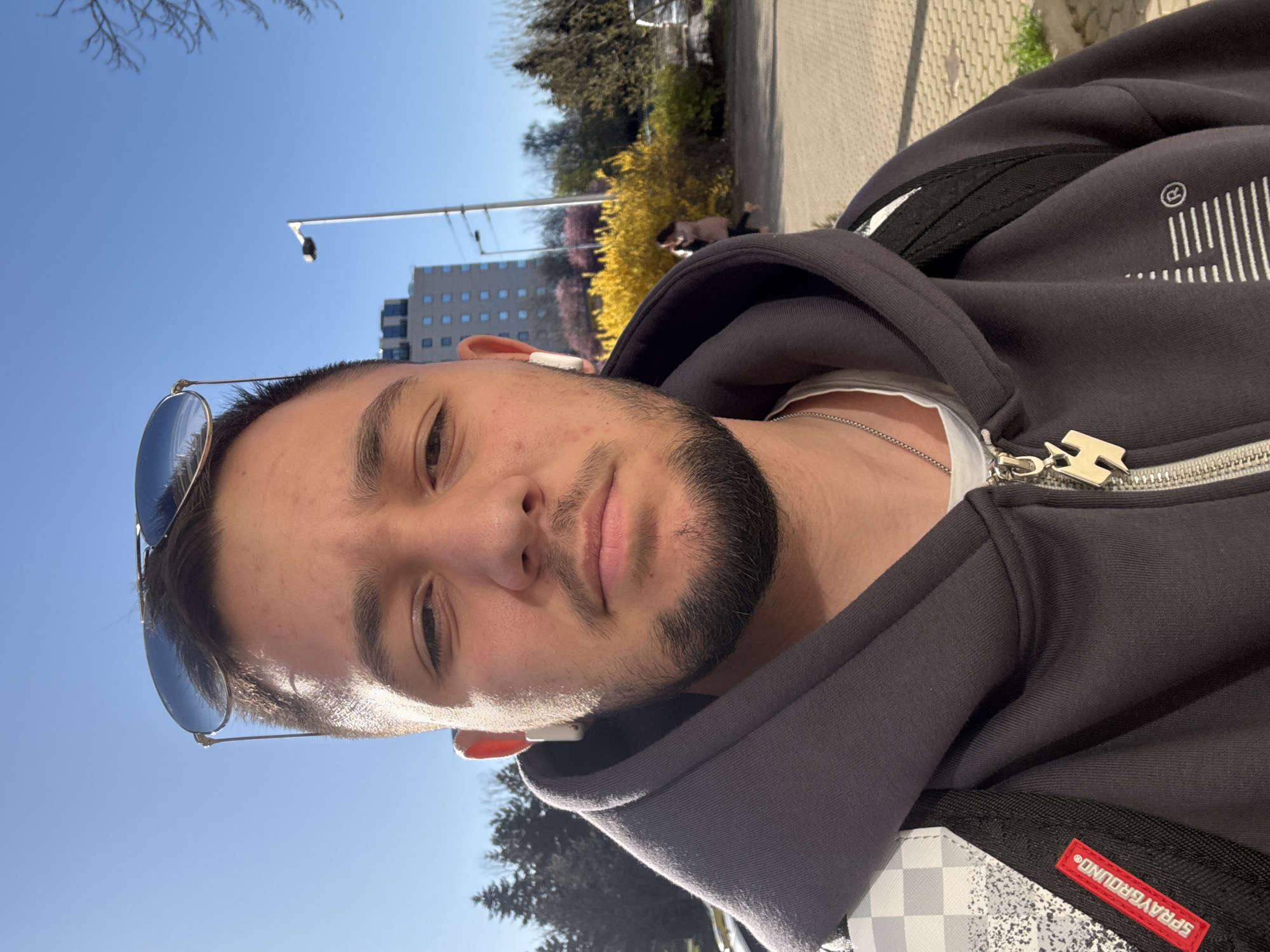 A young man poses for a selfie outdoors in Sofia, Bulgaria. He has a beard and is wearing sunglasses perched atop his head. He wears a gray hooded zip-up sweatshirt, a white t-shirt, and a silver necklace. An earbud is visible in his ear. A backpack is slung over his shoulder with a "SPRAYGROUND" label visible. The background shows a blue sky, bare trees, a light pole, a building with windows, and yellow flowering shrubs. The person is standing on a paved sidewalk next to a green space with trees.