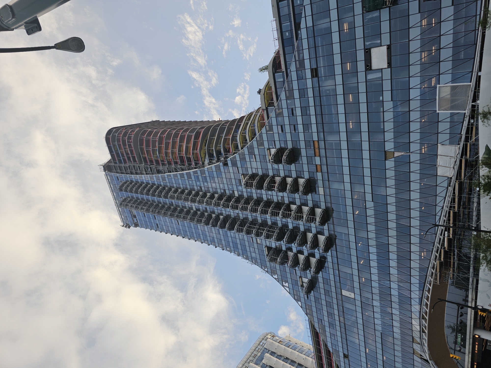 The image shows a modern, glass-facade building under construction in Vancouver, Canada. The building features a distinctive curved design, with the upper floors appearing to twist and taper towards the sky. Several balconies with dark enclosures are visible along the facade of the taller section. The lower portion of the building is characterized by a vast expanse of glass windows, many of which are tinted. In the foreground, scaffolding and construction materials are present, indicating ongoing work. A street light and traffic signals are also visible, suggesting an urban setting. The sky is partly cloudy with a mix of blue sky and white clouds. The architectural style and the presence of construction suggest a contemporary urban development in the city.