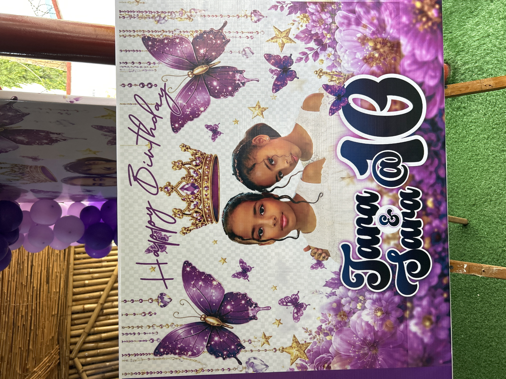 The image depicts a birthday banner for two girls named Tara and Sara who are turning 10. The banner is decorated with purple butterflies, crowns, flowers, and stars. Two young girls, presumably Tara and Sara, are featured prominently on the banner. The banner is displayed outdoors, possibly at an event or party. In the background, bamboo structures and purple balloons are visible. The ground appears to be covered in artificial green grass. The location is identified as Bar Jirgi Summa, Nigeria.