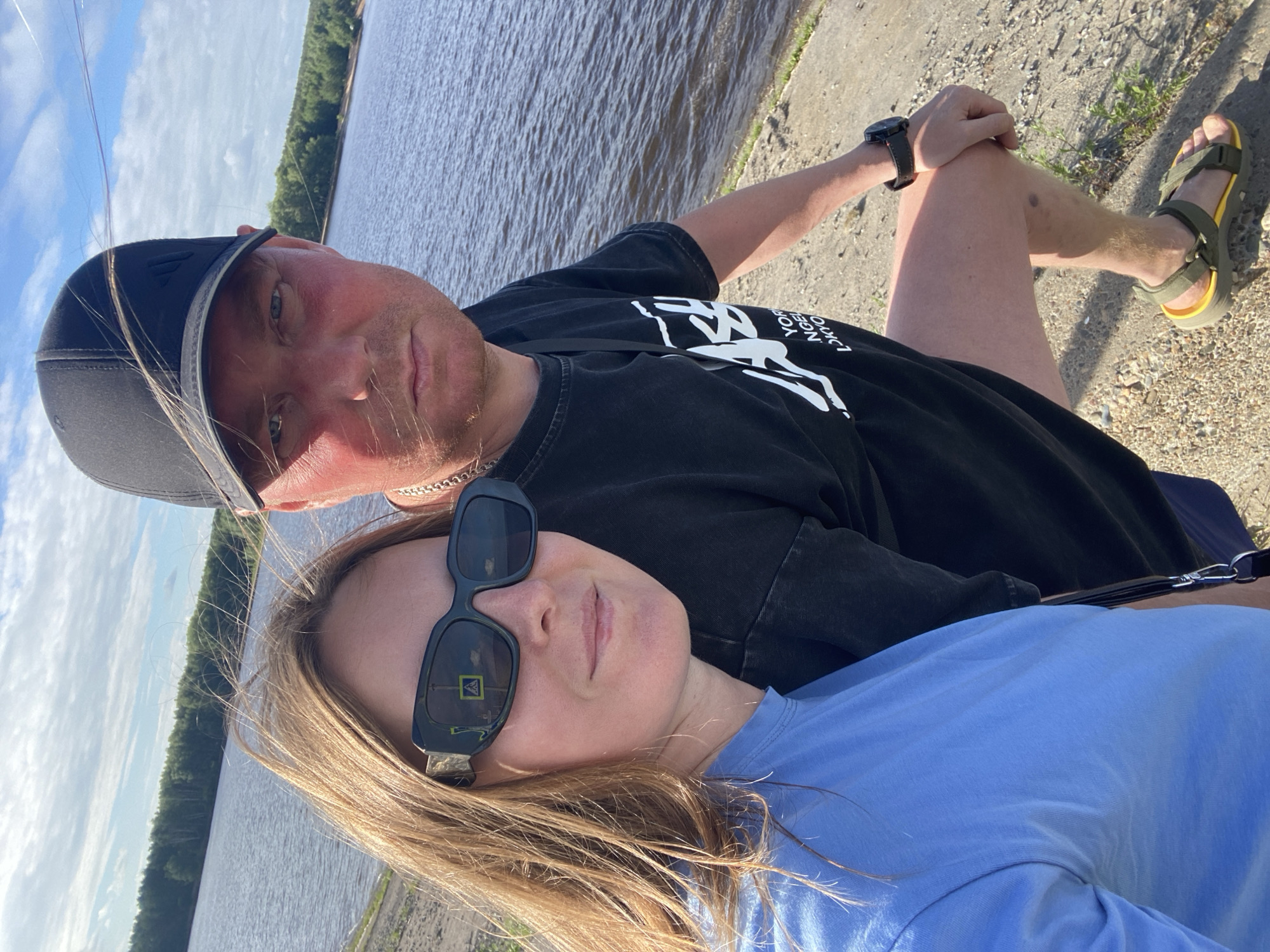 In Noviy, Russia, a man and a woman are taking a selfie by a body of water. The man, wearing a black baseball cap and a black t-shirt, has his arm around the woman. His face is flushed, suggesting sun exposure. The woman, with blonde hair, wears sunglasses and a blue t-shirt. They are positioned on a gravelly bank overlooking the water, with a lush green forest lining the opposite shore under a partly cloudy sky. The image captures a casual outdoor moment.