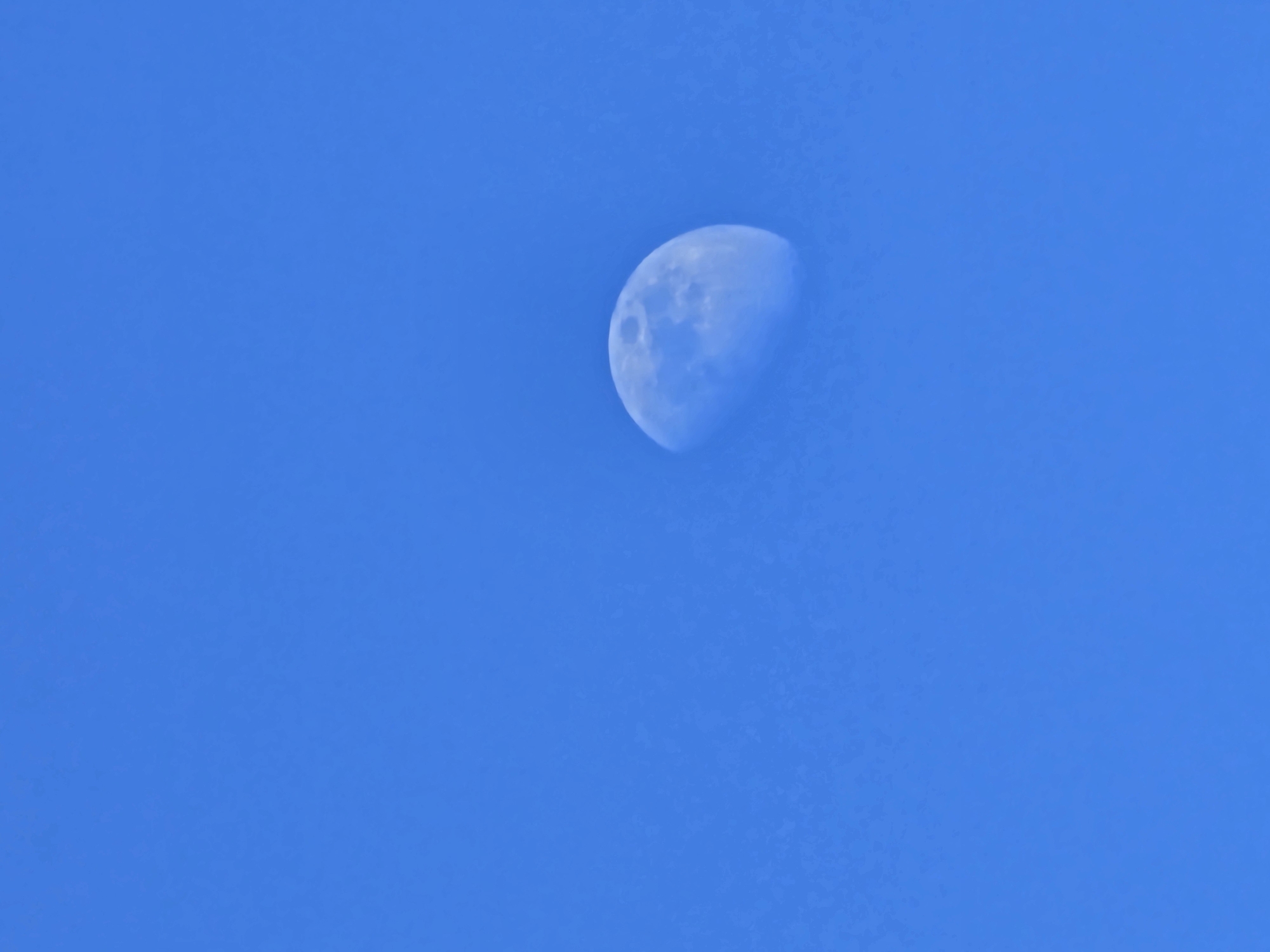 The image depicts a pale, gibbous Moon against a clear, vibrant blue daytime sky. The Moon, appearing in the lower-middle-right portion of the frame, shows discernible surface features including craters and maria, consistent with its illuminated portion. No clouds, terrestrial features, people, or other objects are present. The scene is of the sky above Esch-sur-Alzette, Luxembourg, observed during daylight hours.