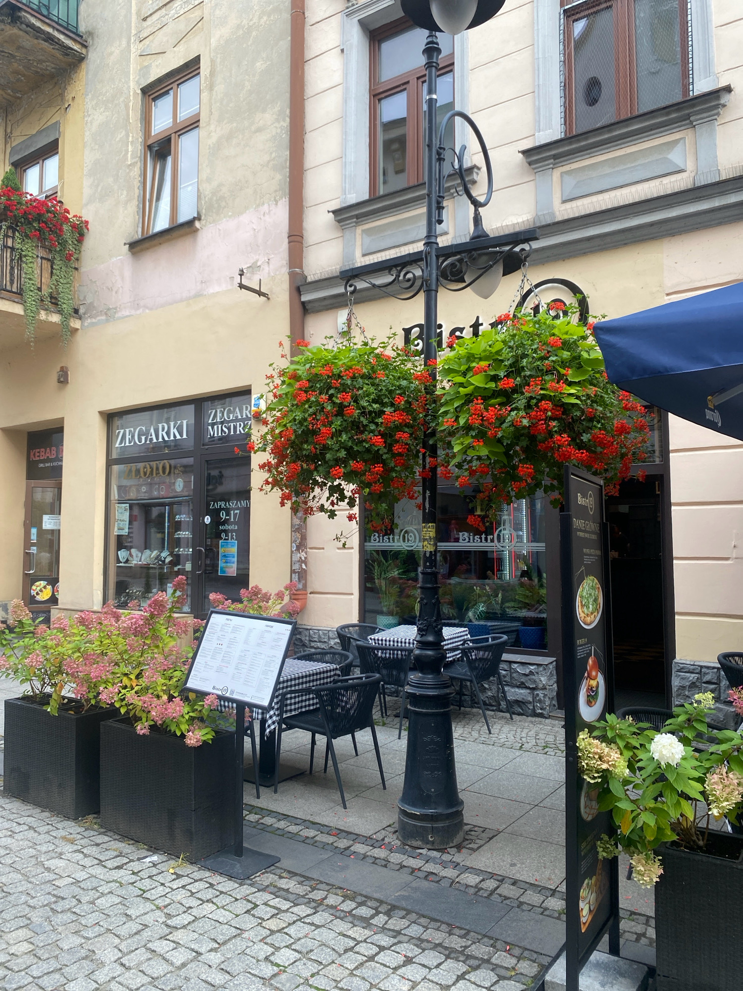 This is a medium-shot, eye-level, outdoor photograph of a street scene in Nowy Sącz, Poland. The architecture suggests a European old town setting, with aged building facades and cobblestone pavement.
On the left, a building has a tan-colored facade with a balcony adorned with red flowers and green hanging plants. Below the balcony, a storefront is visible with a sign indicating "KEBAB GRILL BAR & KUCHNIA". Next to it is another storefront with large glass windows displaying jewelry and a sign reading "ZEGARI MISTRZ ZEGARK I ZLOTO". A menu board stands outside this shop.
In the center of the image, a black lamppost with intricate scrollwork holds two hanging baskets overflowing with vibrant red flowers and green foliage. Adjacent to the lamppost, outdoor seating for a bistro is set up with black chairs and tables covered in black and white checkered cloths. A menu stand is positioned in front of the tables. The bistro's entrance is visible behind the tables, with a sign that appears to say "BISTR 19".
To the right, a dark blue umbrella shades part of the bistro's seating area. Another building facade is visible, with windows and decorative architectural elements. A dark signpost with images of food is placed near the right edge of the frame, partially obscuring the bistro's entrance.
The ground is paved with cobblestones and some stone tiles. The overall atmosphere is that of a quaint European street with businesses and outdoor dining. There are no people visible in the scene.