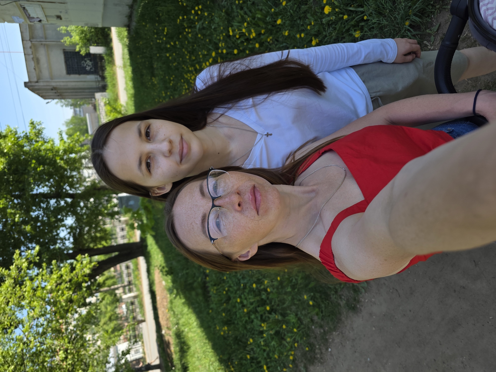 Two women are posing for a selfie outdoors in Chaikovsky, Russia. The woman in the foreground is wearing glasses and a red top. The other woman is wearing a white top. The women are surrounded by grass with yellow flowers. A sidewalk is visible in the foreground. A tree and a building are visible in the background.