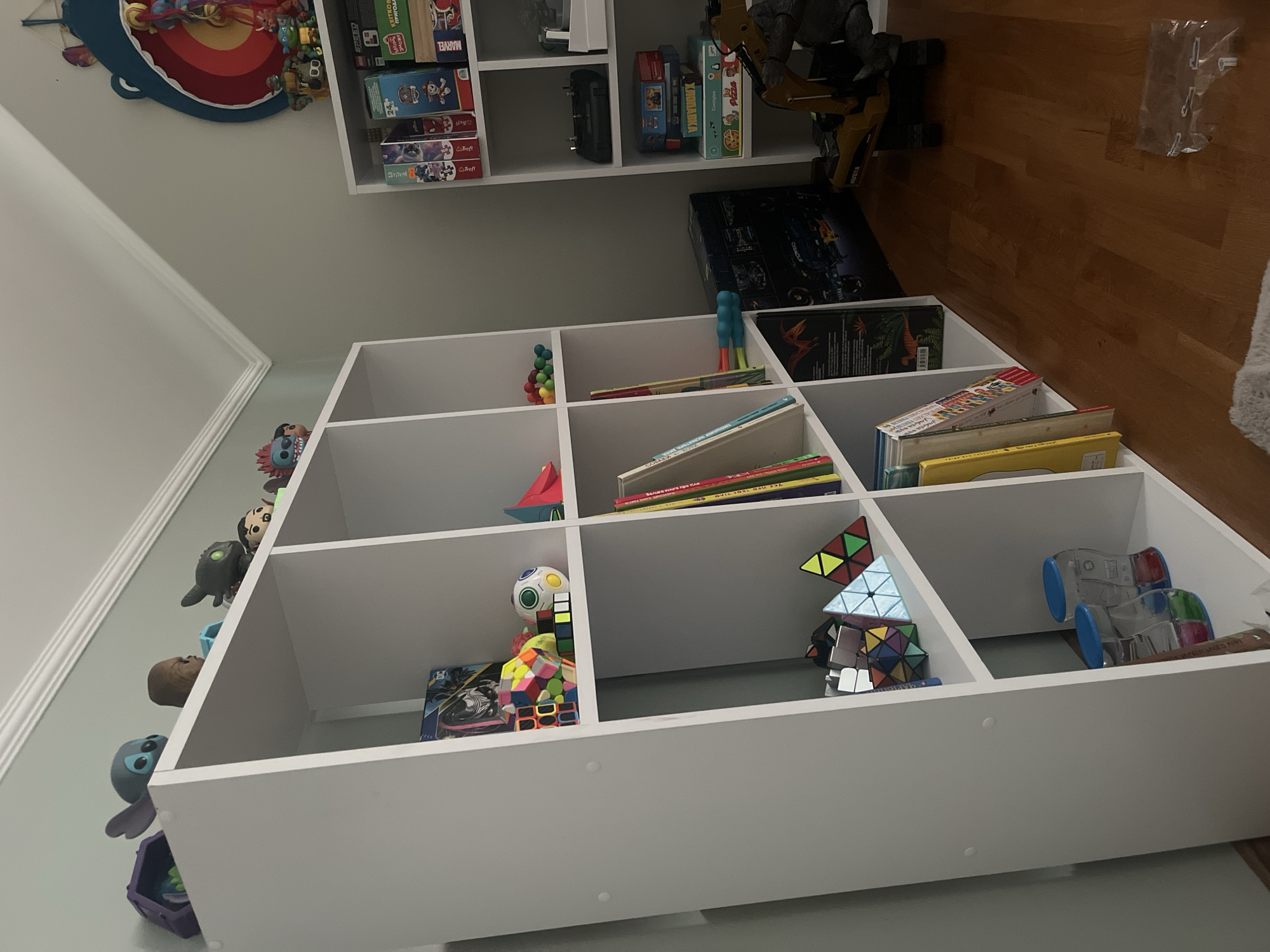 This is a media file featuring a white modular shelving unit filled with various toys and books. The unit is positioned against a light gray wall, with a wooden floor visible on the right side of the frame. The scene appears to be a child's playroom or a storage area for toys.

The shelves contain a diverse collection of items. In the top left, a large, colorful wall decoration resembling a whale's mouth is visible, filled with smaller toys. The shelves below are populated with board games, puzzle boxes, and individual puzzles. Some of the visible text on the boxes includes "MARVEL," "SPEED," and what appears to be text in Ukrainian. There are also figurines of characters like Stitch from Disney's Lilo & Stitch, and other assorted toys in various sizes and colors, including several Rubik's Cubes and other cube-shaped puzzles. Two blue-wheeled toy vehicles are present on the far right shelf.

The lighting suggests it could be daytime, with natural light possibly coming from the left. The setting is indoors, likely a residential space in Onokivtsi, Ukraine, given the location context. There are no people visible in the frame, and the overall impression is one of a well-stocked and organized toy collection.