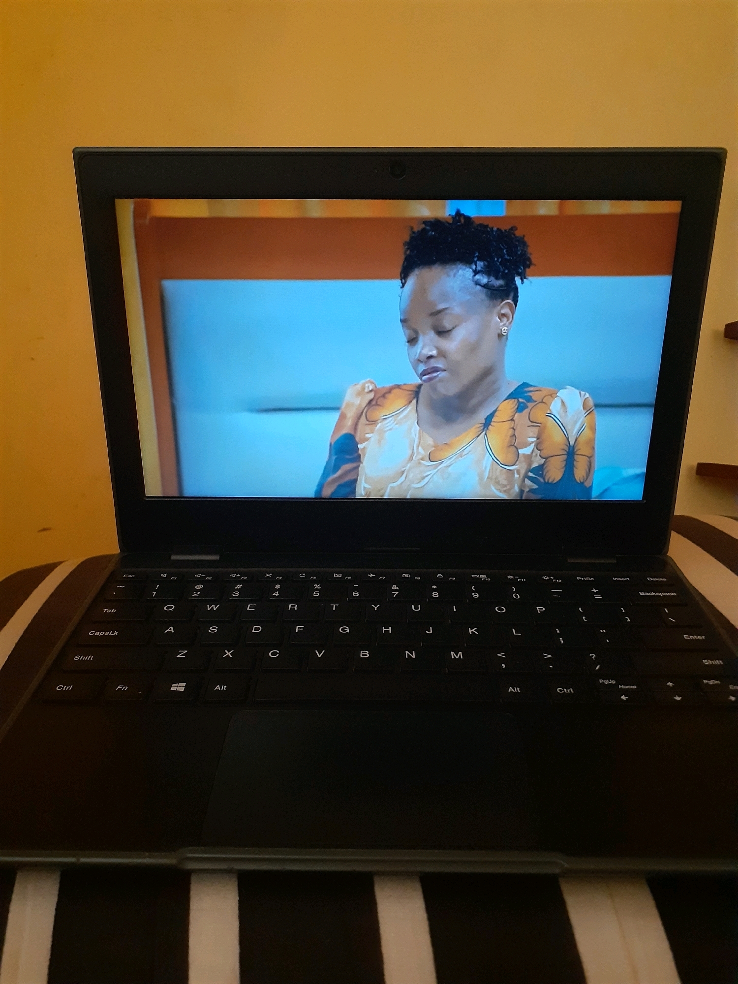 A closed laptop displays a video of a woman with short, styled hair and a light-skinned complexion. She is wearing a garment with a pattern of orange and blue butterfly-like shapes. The background of the video is a light blue. The laptop is positioned on a striped surface, likely a bed, with a yellow wall in the background. This scene is located in Nyanguge, Tanzania.