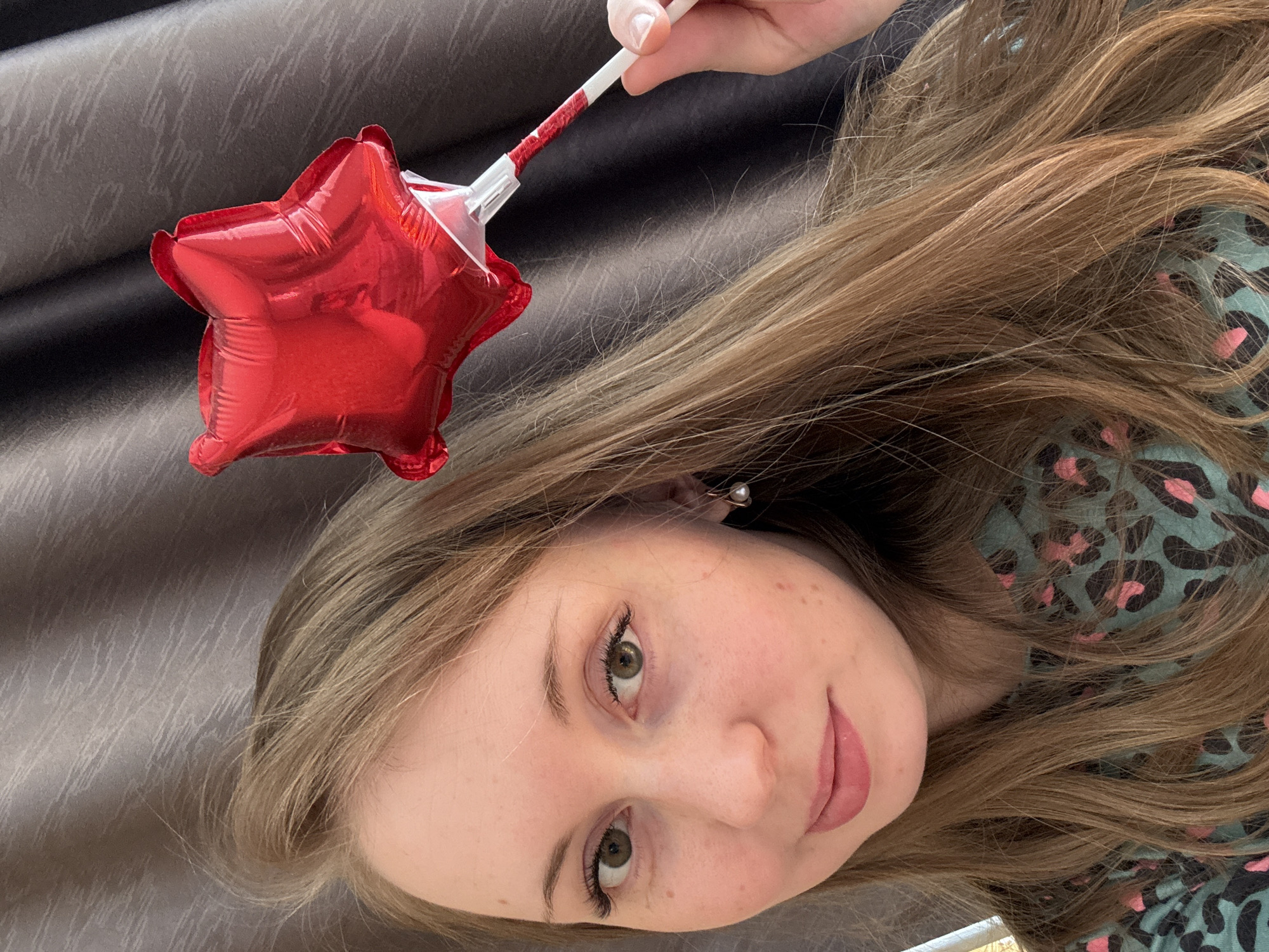 An overhead portrait shows a young person with long, light brown hair, angled looking up towards the camera. The image is rotated 90 degrees clockwise. The person has fair skin, visible freckles, and light green or hazel eyes. A subtle, natural-looking lip color is applied. They are wearing a teal-colored top with a black and pink leopard print pattern. A small, pearl-like earring is visible in their right ear.
The person's right hand (upper part of the image) holds a small, inflatable red star-shaped balloon attached to a white stick adorned with red glitter. The background consists of a dark, textured fabric, possibly a curtain or wall covering, creating an indoor setting. There is no discernible specific activity other than posing for the photograph. The person's expression is neutral, with a slight hint of a smile. No text is visible in the image.