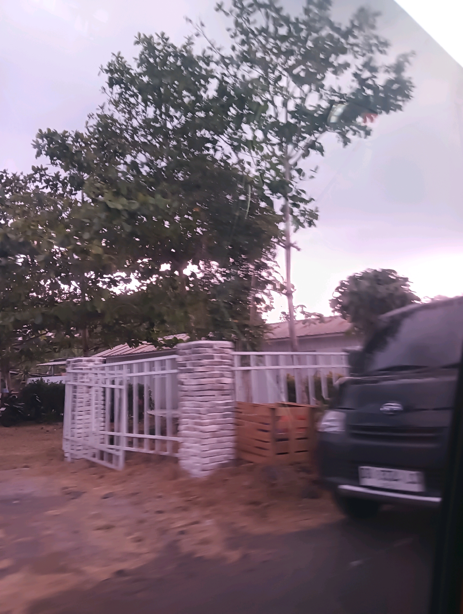 The image depicts a street scene with a white metal fence and gate, a brick pillar, and a wooden crate. A dark-colored vehicle is parked on the right side of the frame. Trees with green foliage are visible in the background, and the sky is overcast with a purple hue. The overall impression is that of a residential or semi-commercial area, possibly in a tropical or subtropical climate given the vegetation. Without additional context or visible landmarks, it is not possible to definitively determine the exact location within Tomohon, Indonesia.
