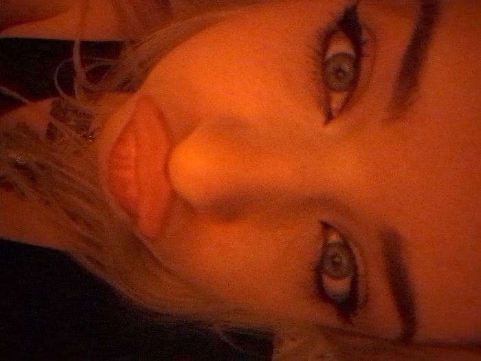 This image captures a close-up, vertically oriented view of a human face, located in Area D (Elaho/Garibaldi), Canada. The subject, an individual with light-colored eyes, features prominent dark winged eyeliner and defined dark, arched eyebrows. The entire face is uniformly illuminated by a dominant orange-red light source. The lips are full, reddish, and intentionally formed into a puckered or 'pout' expression. Strands of light-colored hair are visible framing the lower face. A dark, slender object, potentially a necklace or chain, is faintly visible below the chin against an indistinguishable dark background. No other individuals, objects, or environmental details are present within the frame.