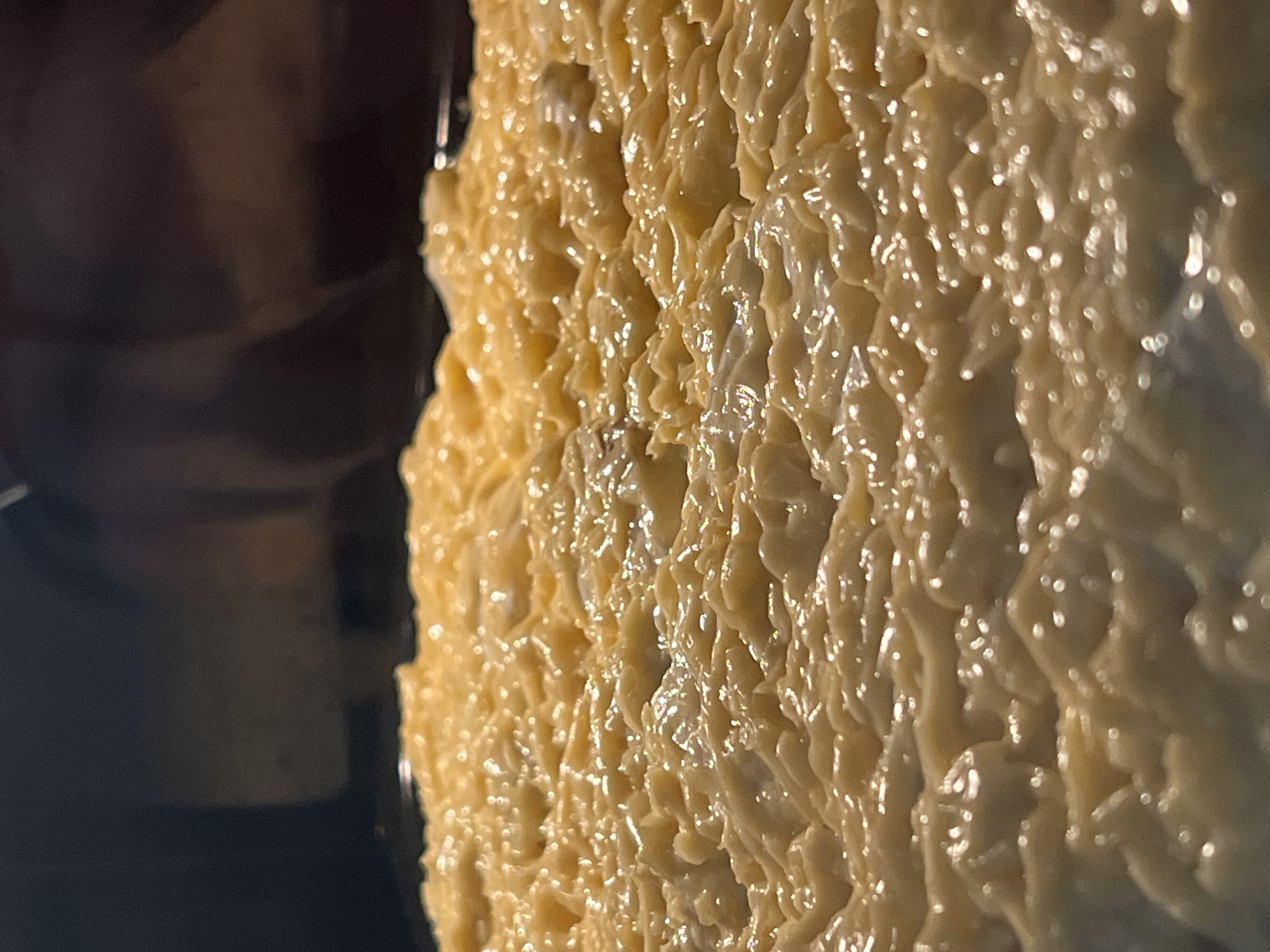 This is a close-up image of a food item, possibly a cake or dessert, covered in a thick, textured, glossy, light-yellow icing or frosting. The texture of the frosting appears uneven, with numerous small peaks and valleys, reflecting light. To the left of the frosted area is a dark, blurred area which could be the background or the rest of the dessert. The location and city are unknown.
