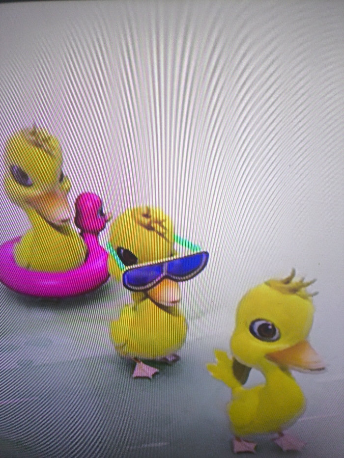Three yellow cartoon ducklings are shown. One wears sunglasses, another has a pink floatation device around its body, and the third is standing with its wing raised. The ducks have pink feet and orange beaks. The backdrop is a light grey with faint vertical lines. The image is possibly from a television or monitor.