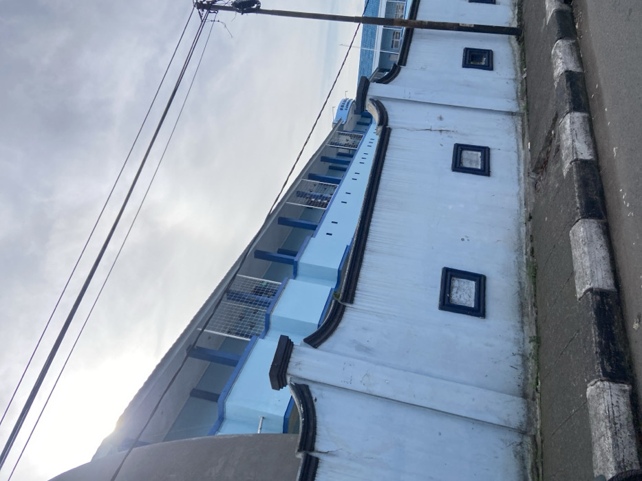 The image shows a blue building with a white fence, a sidewalk, and a street in Margahayu, Indonesia. The building is made of concrete and has a flat roof. The fence is made of concrete and is painted white. The sidewalk is made of concrete and has a black and white checkerboard pattern. The street is paved with asphalt. The sun is shining brightly. The building has multiple rows of windows with black frames and blue trim, and the roof has a blue trim as well. There are two power lines above the building. One of the power lines has an attached utility pole that reaches into the frame of the image. There are no people in the image. The building appears to be abandoned.