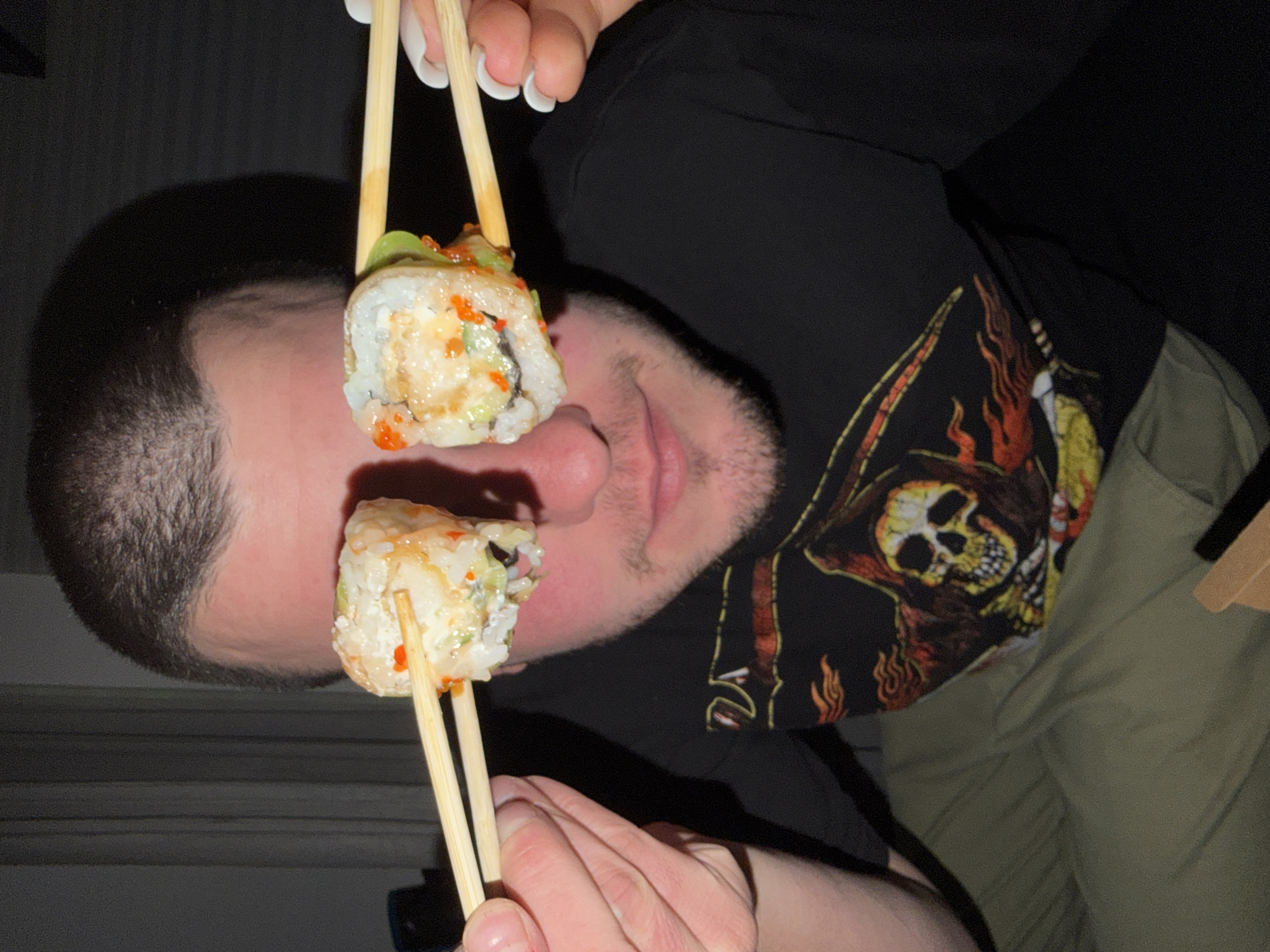 A man holds sushi rolls on chopsticks in front of his face. He wears a black t-shirt with a skull graphic and khaki pants. The location is indeterminate; there is no evidence to suggest it is Varna, Bulgaria.