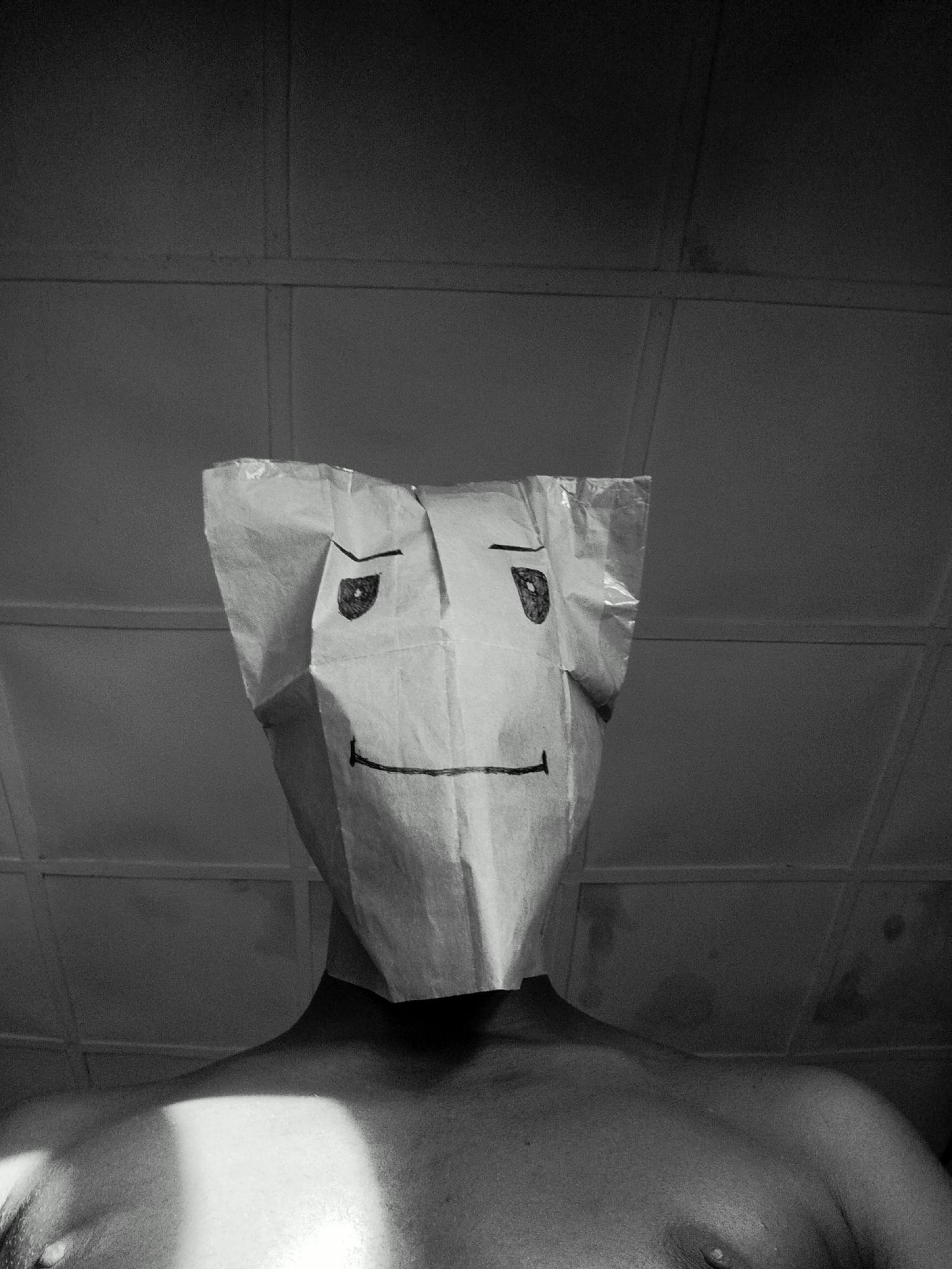 A black and white, low-angle medium shot depicts a shirtless person with a paper bag over their head. The bag has a crudely drawn face with slanted eyebrows and a simple, horizontal line for a mouth. The person's neck and upper chest are visible. The background shows a ceiling with a tiled or paneled design. There is no information to suggest the image was taken in Keffi, Nigeria.