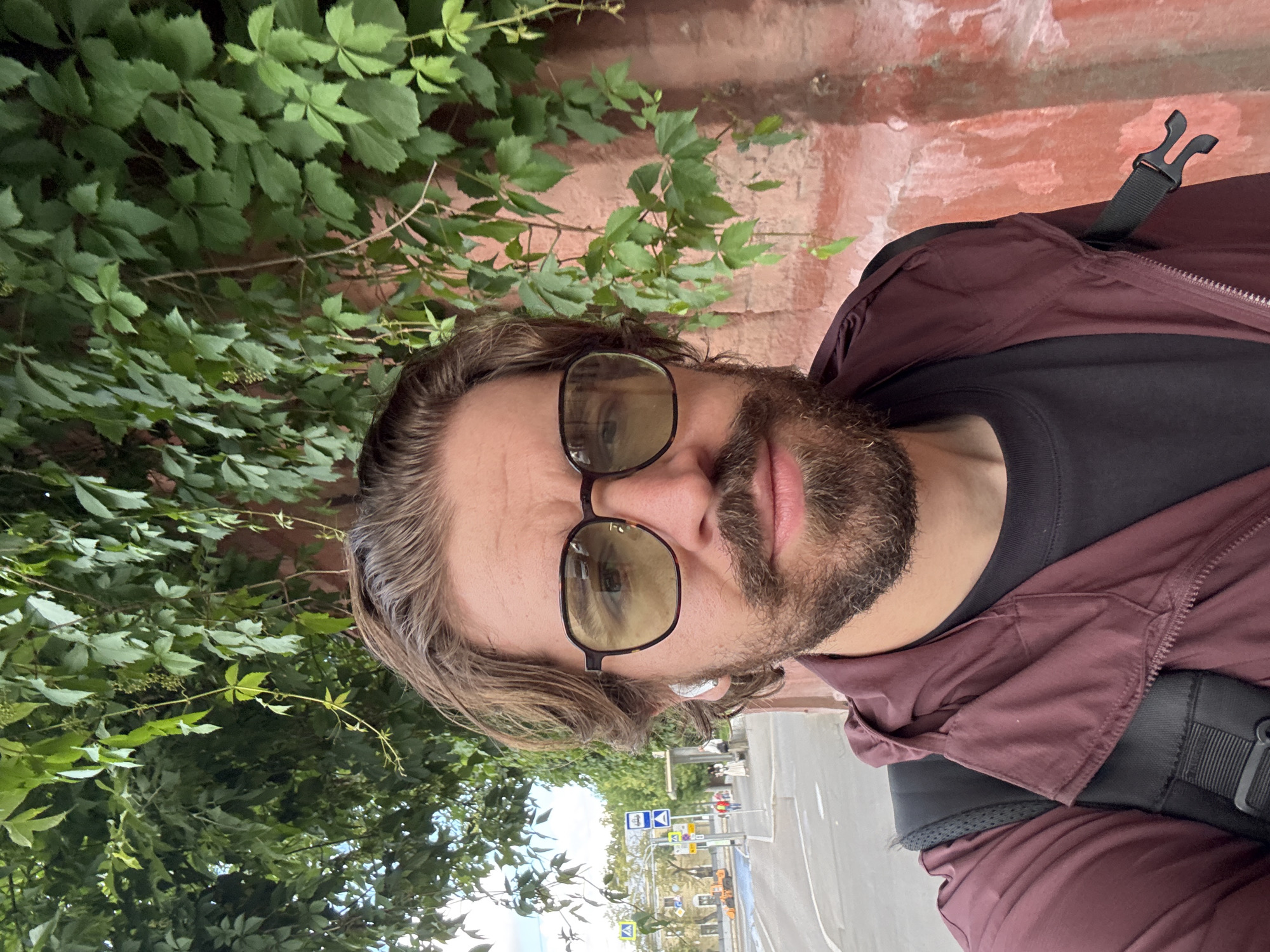 A man is taking a selfie outdoors, likely in Moscow, Russia, given the visible road signs. He is wearing sunglasses, a dark t-shirt, and a burgundy jacket. He has brown hair and a beard. A backpack strap is visible over his shoulder. He appears to be standing next to a brick wall covered in green vines. In the background, there is a street with traffic and various road signs, suggesting it is daytime. The weather seems overcast. The image is oriented vertically, with the man's head towards the top of the frame and his body extending downwards.