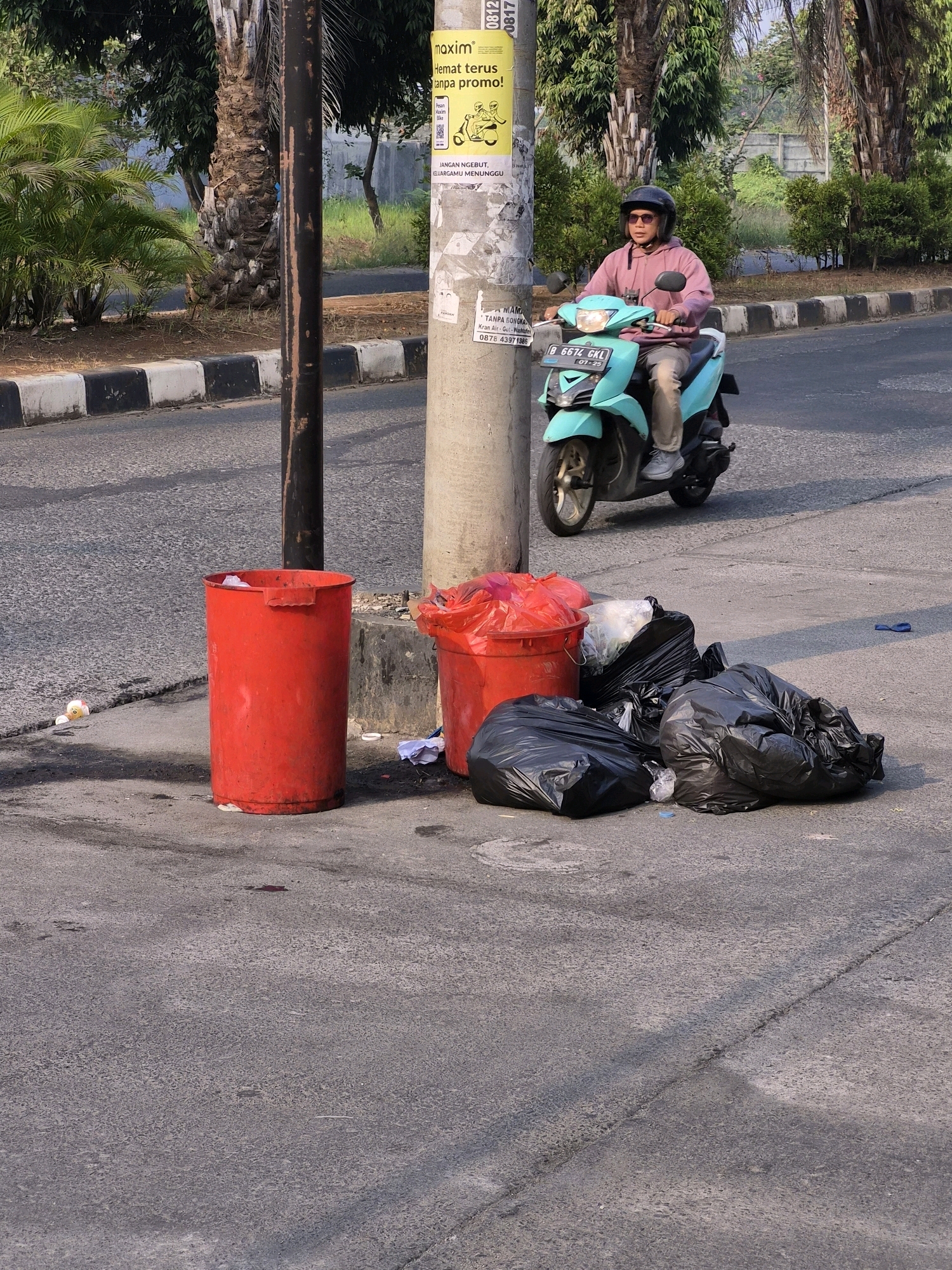 A medium shot captures a scene on a roadside in Wanajaya, Indonesia, under bright daylight. In the foreground, a red cylindrical trash can stands to the left, filled with some white debris. To its right, several black trash bags are piled on the pavement, with a red plastic bag on top of a white plastic bag peeking out from behind the trash can. Two concrete poles are visible behind the trash and bags. In the mid-ground, a person wearing a pink hoodie, sunglasses, and a black helmet rides a turquoise scooter. The scooter has a license plate that reads "B 6674 GKL". The person appears to be looking forward. The background features a street with a black and white striped curb, trees, and a light-colored wall.