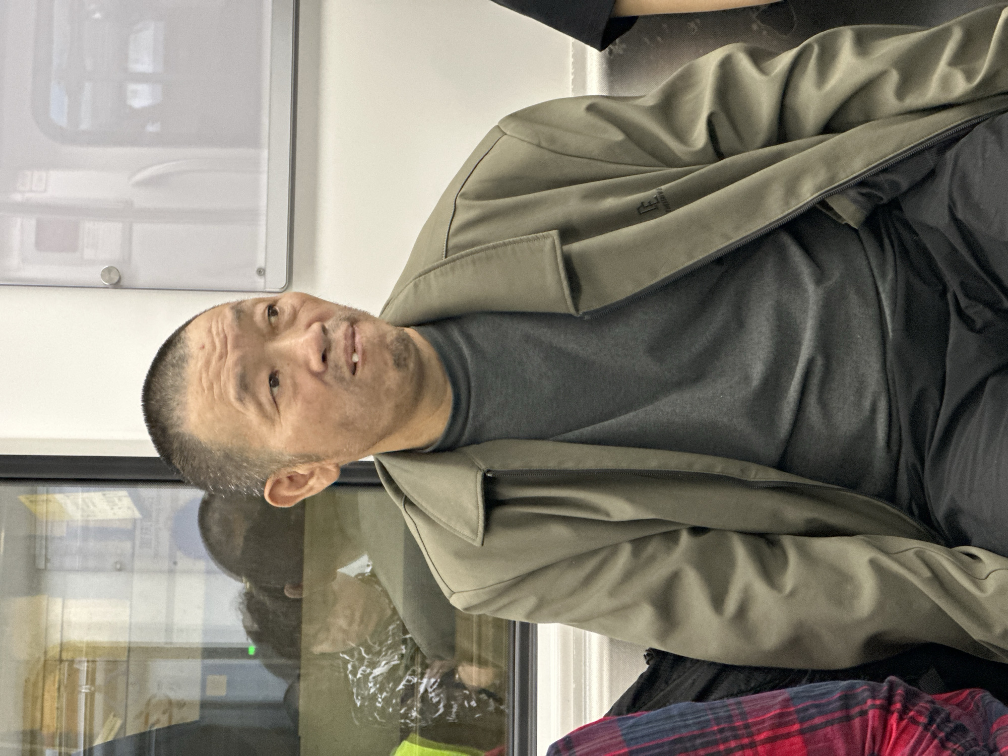 A medium shot depicts an Asian male with short, dark hair, facing the camera. He is wearing a gray shirt and an olive green jacket. The background reveals what appears to be a window reflecting a blurry cityscape. This suggests that the location may be inside a bus or train. A plaid shirt is visible at the bottom. The exact city of Unknown, Unknown, cannot be determined from this single frame.
