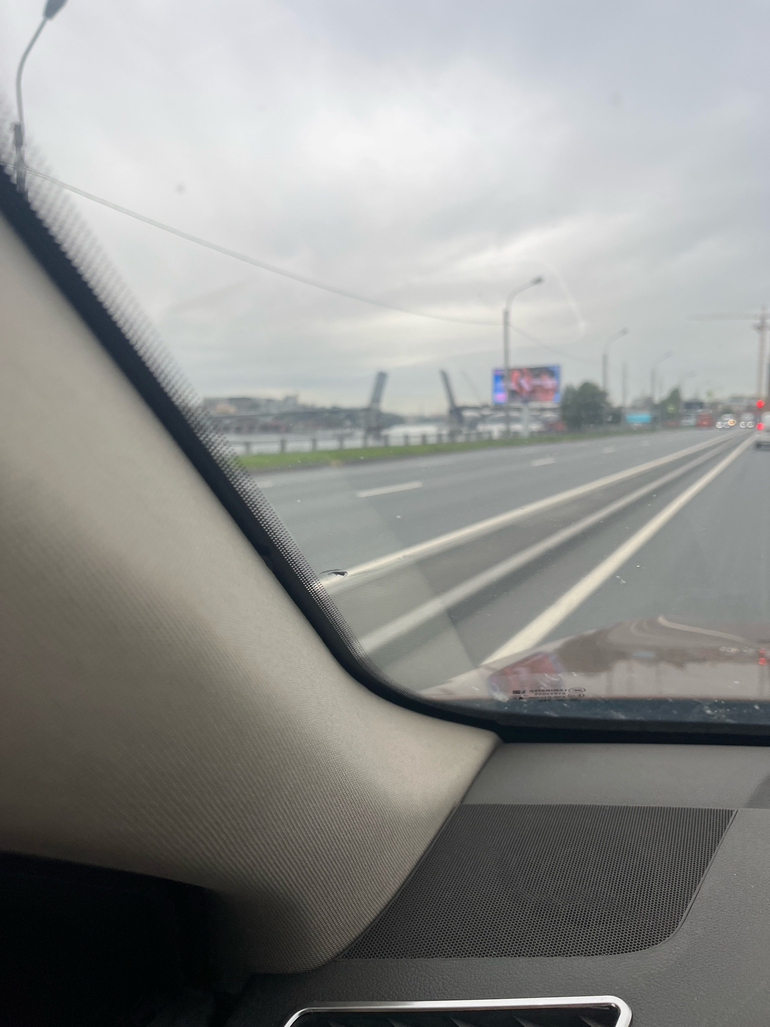 The image is taken from inside a vehicle looking out through the windshield on an overcast day. The foreground shows the interior of the car, including a textured dashboard and a speaker grille. The middle ground shows a wet road with white lane markings and a grassy median. In the background, there are industrial structures, a large billboard with an advertisement, streetlights, and distant buildings. The scene is consistent with a view of the Gulf of Finland or a large body of water adjacent to the city, potentially near the St. Petersburg Ring Road (KAD) or the Western High-Speed Diameter, given the presence of industrial infrastructure and a wide highway. The overall atmosphere is somewhat gloomy due to the cloudy weather. There are no people visible in the image.