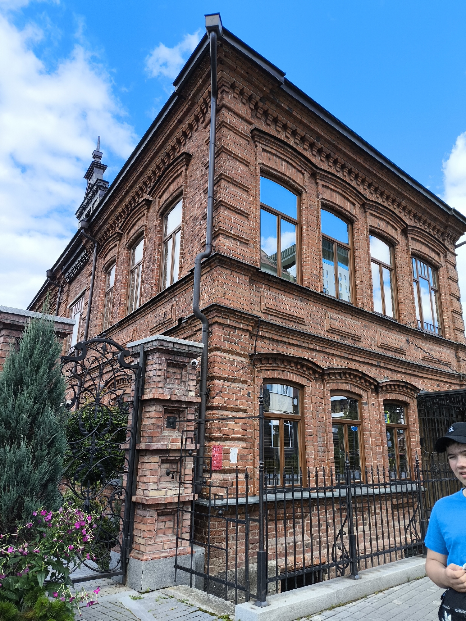 A full, eye-level shot captures the exterior of a two-story, red-brick building with a dark roof. The building is situated in Yekaterinburg, Russia, and features ornate architectural details, including decorative brickwork along the eaves and around the arched windows. Several large, multi-paned windows reflect the bright blue sky and scattered white clouds. A black metal fence with intricate scrollwork encloses the property, and a young person wearing a blue t-shirt and a black baseball cap is partially visible on the right side of the frame, facing towards the building. A tall evergreen tree and some flowering plants are visible to the left, near the entrance. The ground is paved with light-colored stones, and a drainpipe runs vertically down the side of the building.