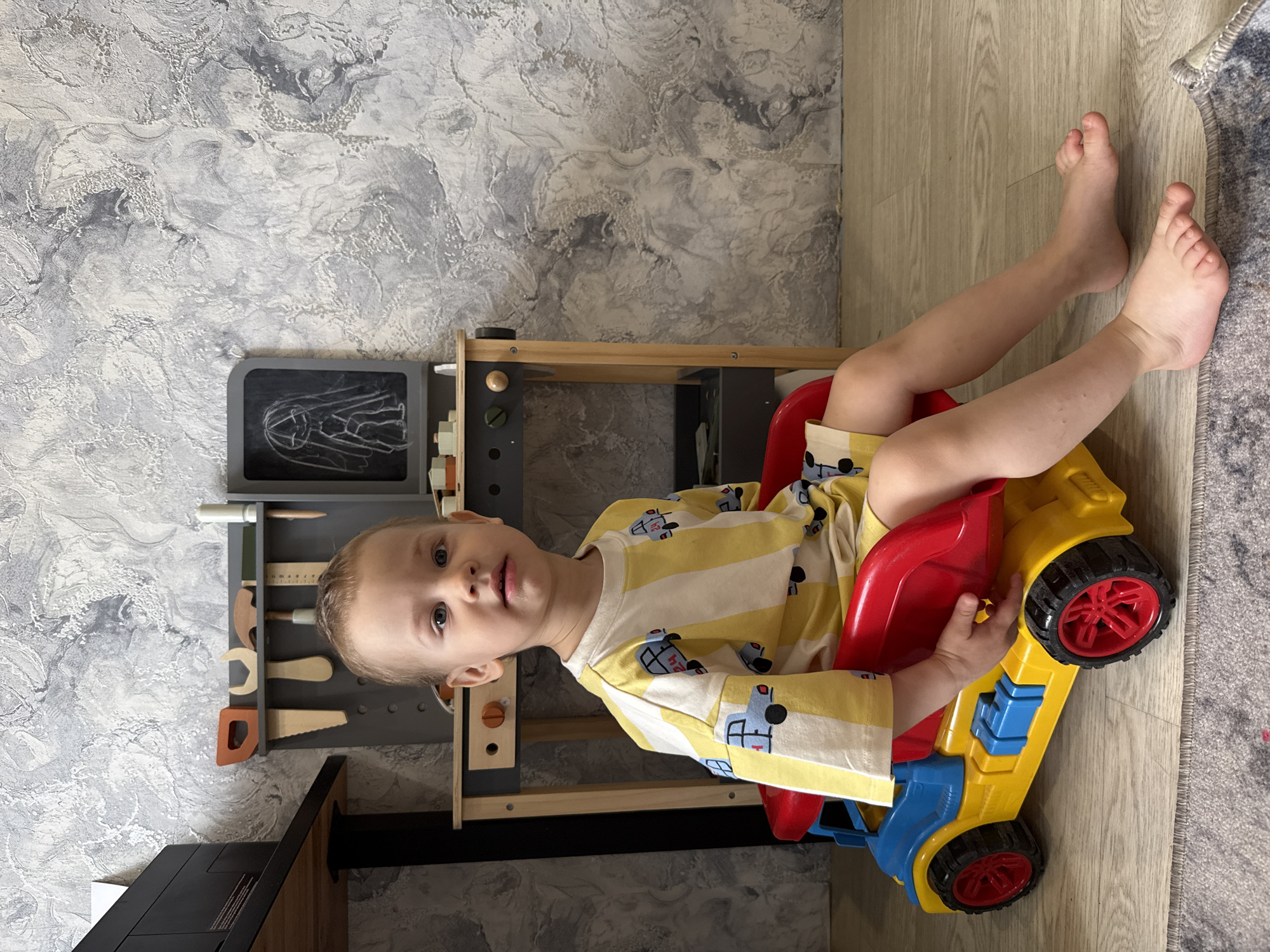 The image captures a young child seated in a colorful toy car indoors, likely in a home in Rusanovo, Russia. The child, appearing to be a toddler or preschooler with light hair and fair skin, is wearing a yellow and white striped long-sleeve shirt patterned with blue cars, and light-colored shorts. They are barefoot, with their legs extended on a light wooden laminate floor that borders a gray patterned rug.
The toy car is large, predominantly yellow, with a red seat and blue structural accents, featuring red wheels. The child is looking towards the camera with a neutral, slightly inquisitive expression. Behind the child and to their left is a wooden toy workbench or tool station, colored in gray and natural wood tones. Various wooden toy tools, including a wrench, hammer, saw, ruler, and what appears to be a screwdriver, are neatly hung on its surface. Integrated into the workbench is a small gray chalkboard displaying a chalk drawing of a figure, possibly a simplified human or skeleton.
The background wall is covered with a textured wallpaper featuring a marbled pattern in shades of gray and white. The lighting suggests it is daytime. The scene depicts a playful, domestic environment, likely a child's room or play area. There is no discernible text visible in the image.