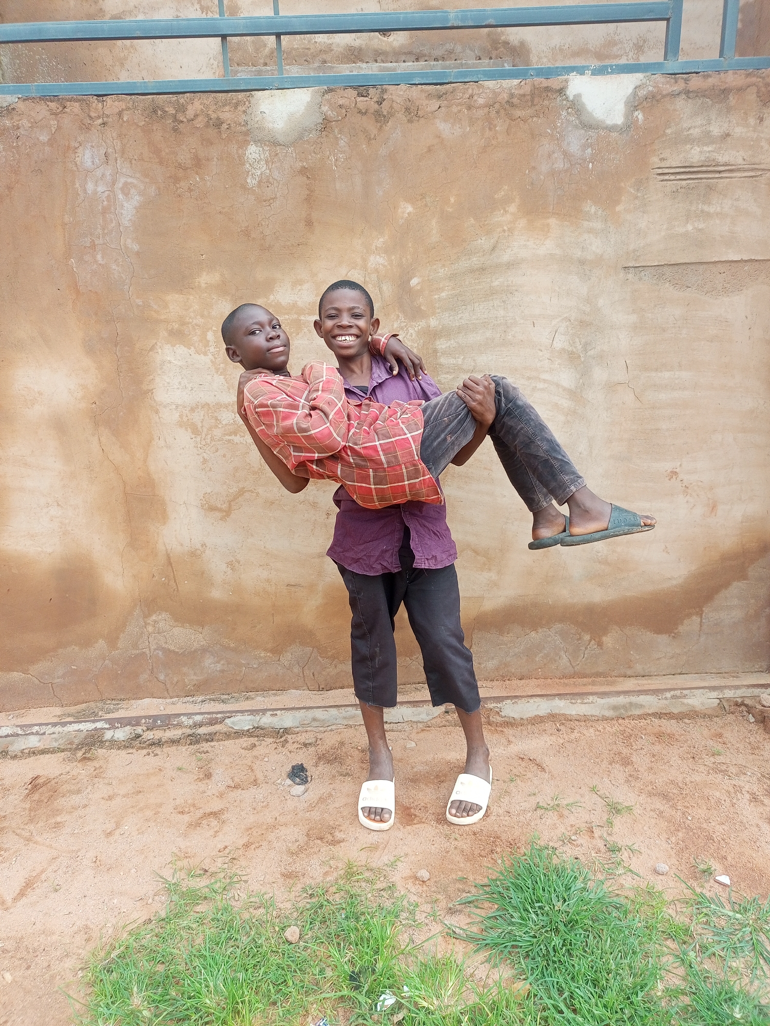Two young boys are in the foreground of the image, standing in front of a textured, light brown wall. The boy on the left, wearing a red and white checkered shirt and dark pants, is being carried horizontally in the arms of the boy on the right. The boy on the right is wearing a purple shirt and dark pants and is smiling broadly at the camera. Both boys are wearing sandals. The ground in front of them is sandy with patches of green grass. Above the wall, a metal railing is visible against a lighter background. The scene appears to be outdoors in an urban or semi-urban environment.