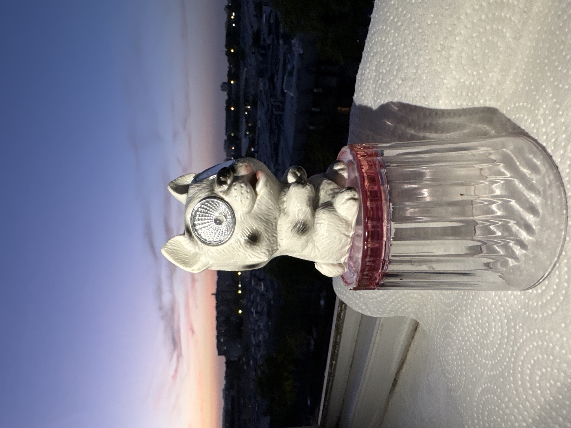 A close-up, eye-level shot captures a white, stylized dog figurine perched atop a clear plastic cup with a pink rim. The figurine has large, round, silver solar panel eyes and dark spots on its body. The cup rests on a white, textured surface, possibly a paper towel. In the background, the sky displays a twilight gradient of deep blue to soft pink, with a hazy cityscape silhouetted against it. Buildings and scattered lights are visible in the distance. The scene is set outdoors, likely on a windowsill or balcony, overlooking a residential area.