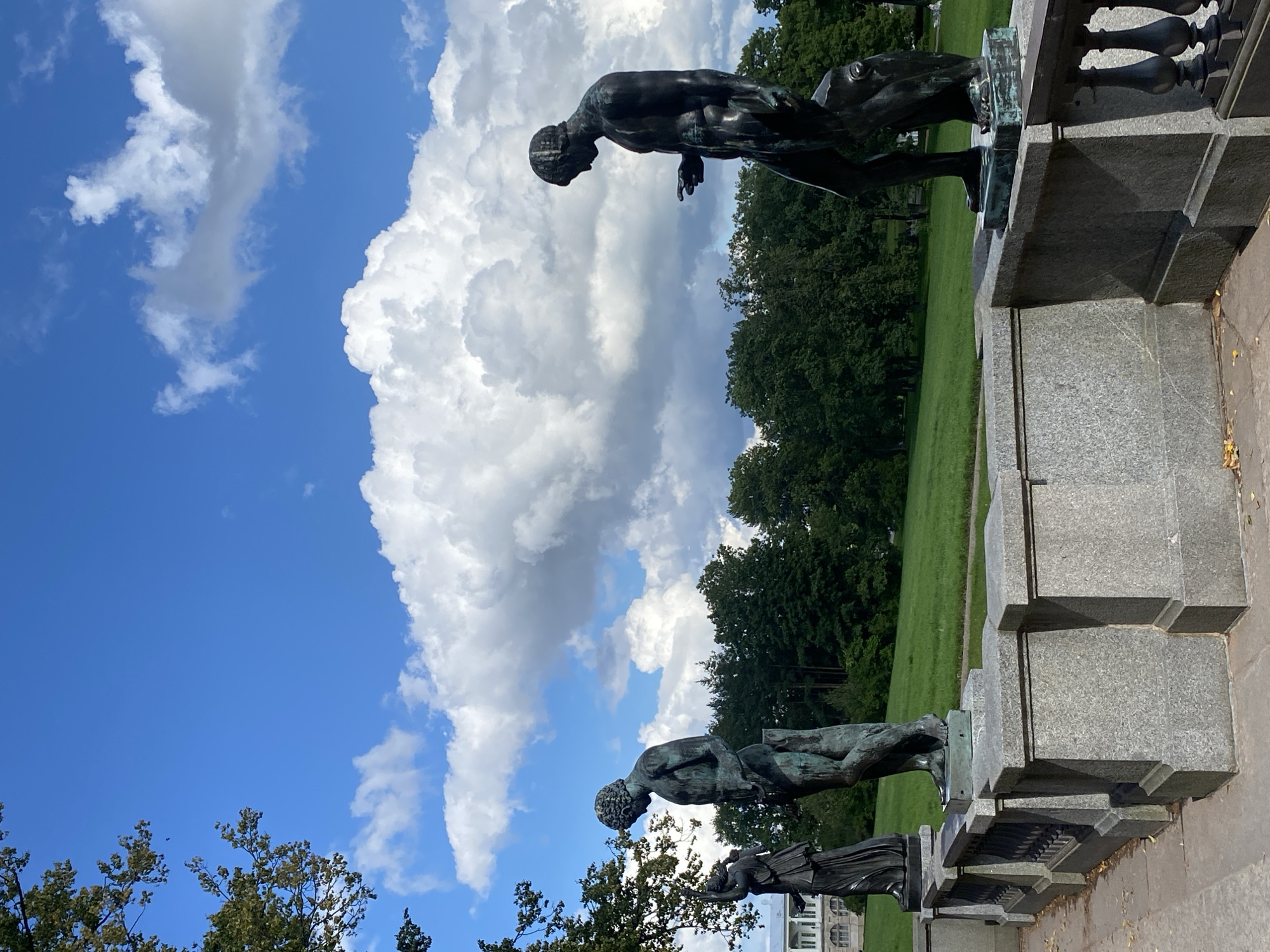 The image depicts a section of the Catherine Park in Pushkin, Russia, specifically a terrace with bronze statues overlooking a manicured lawn and a treeline. Three full-body bronze statues of male figures stand on pedestals along a stone balustrade. The statue on the far left is slightly obscured by a tree branch. The middle statue is a standing male figure, and the statue on the right is also a standing male figure, appearing to point with one hand. The sky above is a vibrant blue, punctuated by large, fluffy white clouds. The background consists of a lush green lawn and a dense forest of mature trees. A portion of a white classical building with columns is visible to the far left of the frame. The lighting suggests it is daytime with bright sunlight.
