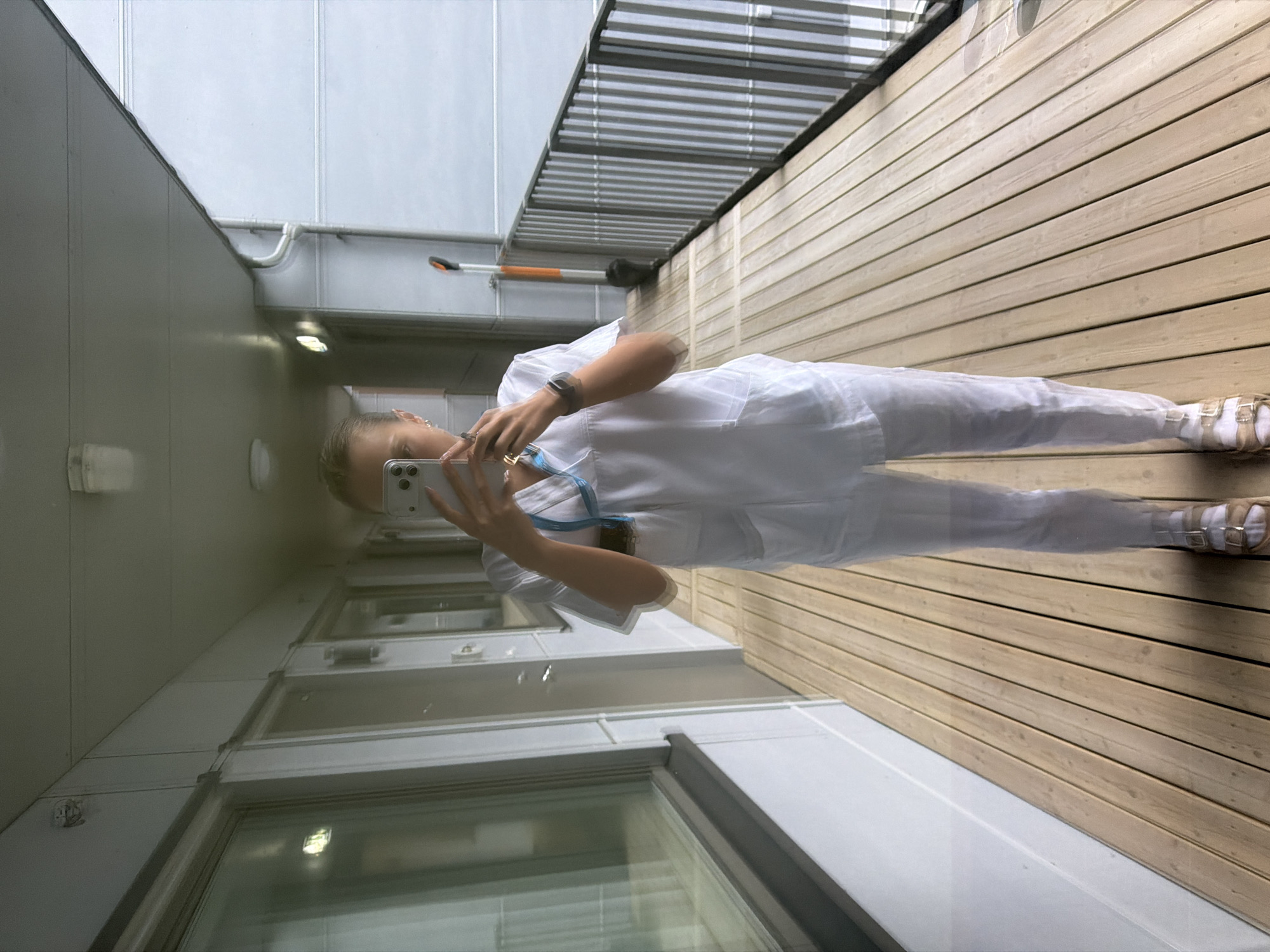 A person, appearing to be a woman with blonde hair pulled back, stands on a wooden deck, holding a smartphone to take a selfie in what appears to be a reflective surface. She is wearing a white scrub suit with a blue stethoscope draped around her neck. She has on white sandals with gold buckles. The environment is an outdoor hallway with doors to rooms on the left and a metal railing on the right overlooking a gray exterior. A broom with an orange handle leans against the railing in the background. The deck is made of light-colored wooden planks. The scene is likely in a residential or institutional building in Harstad, Norway, as indicated by the architectural style and exterior view.
