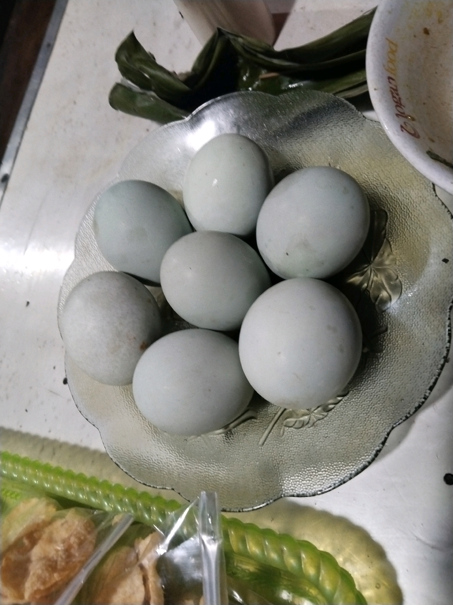 A close-up, overhead shot shows a clear, textured glass bowl filled with seven light blue-green eggs. The bowl is positioned on a light-colored, speckled surface that appears to be a table. Behind the bowl, a few dark green leaves are visible, as well as a white bowl with some red lettering. In the foreground, a clear plastic bag contains what looks like fried snacks and a long, green, ribbed vegetable. The lighting suggests an indoor setting, possibly a kitchen. The image does not contain any identifiable landmarks or features that would specifically place it in the city of Karanganyar, Indonesia.