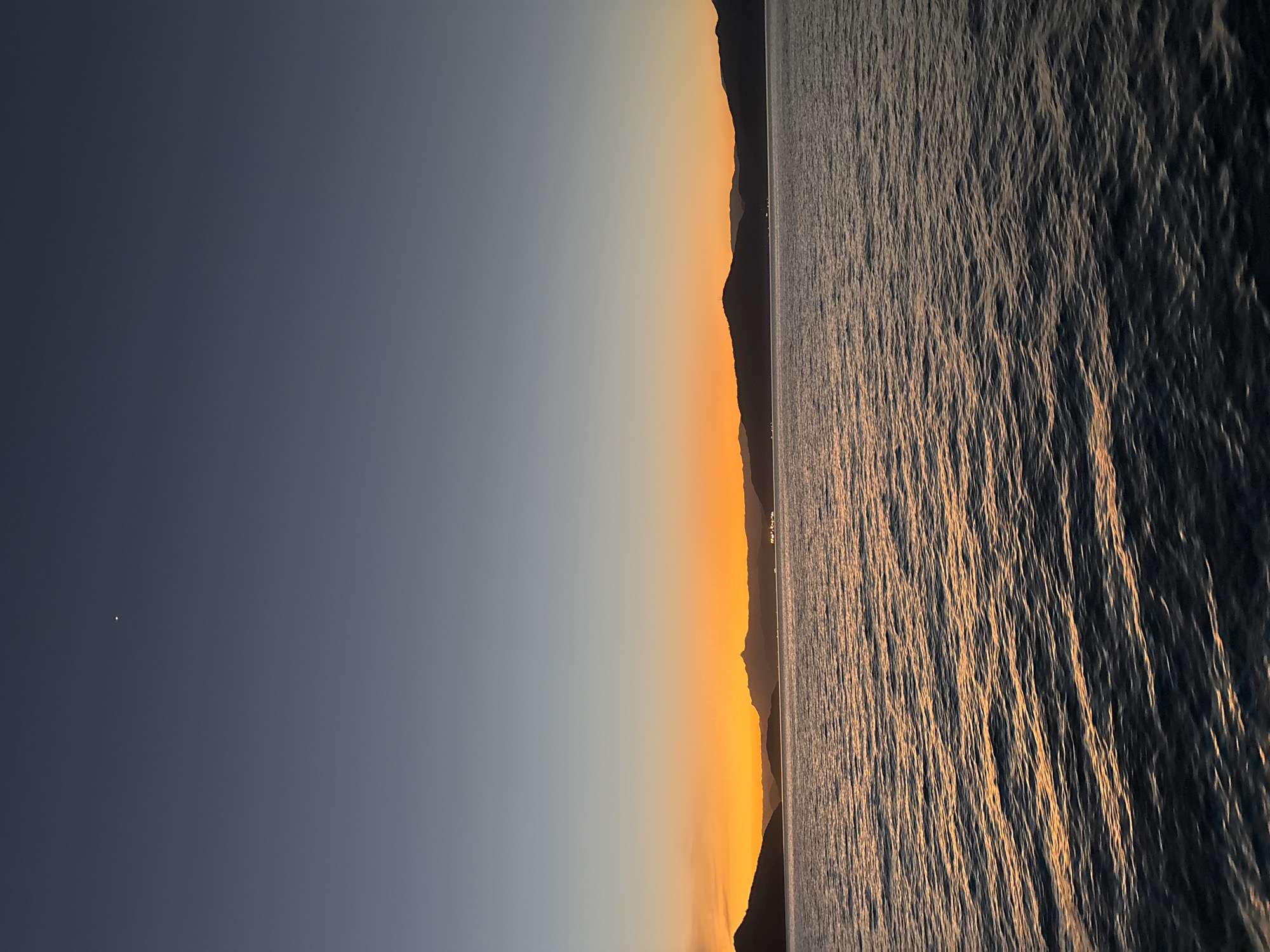 The image depicts a serene sunset over a body of water, likely a bay or strait, with silhouetted mountains forming the horizon. The sky transitions from a deep blue at the top to vibrant shades of orange and yellow near the horizon, indicating the sun has recently set or is about to rise. A single, small bright point, possibly a star or planet, is visible in the upper portion of the sky. The water's surface is gently rippled, reflecting the warm colors of the sky. The scene is devoid of any people or visible man-made structures except for a faint suggestion of lights on the distant shore. Given the characteristic geography of Angra dos Reis, Brazil, this scene is likely captured from a viewpoint within or near the city, overlooking the Bay of Ilha Grande, with the surrounding mountains of the Serra do Mar forming the dramatic silhouette.