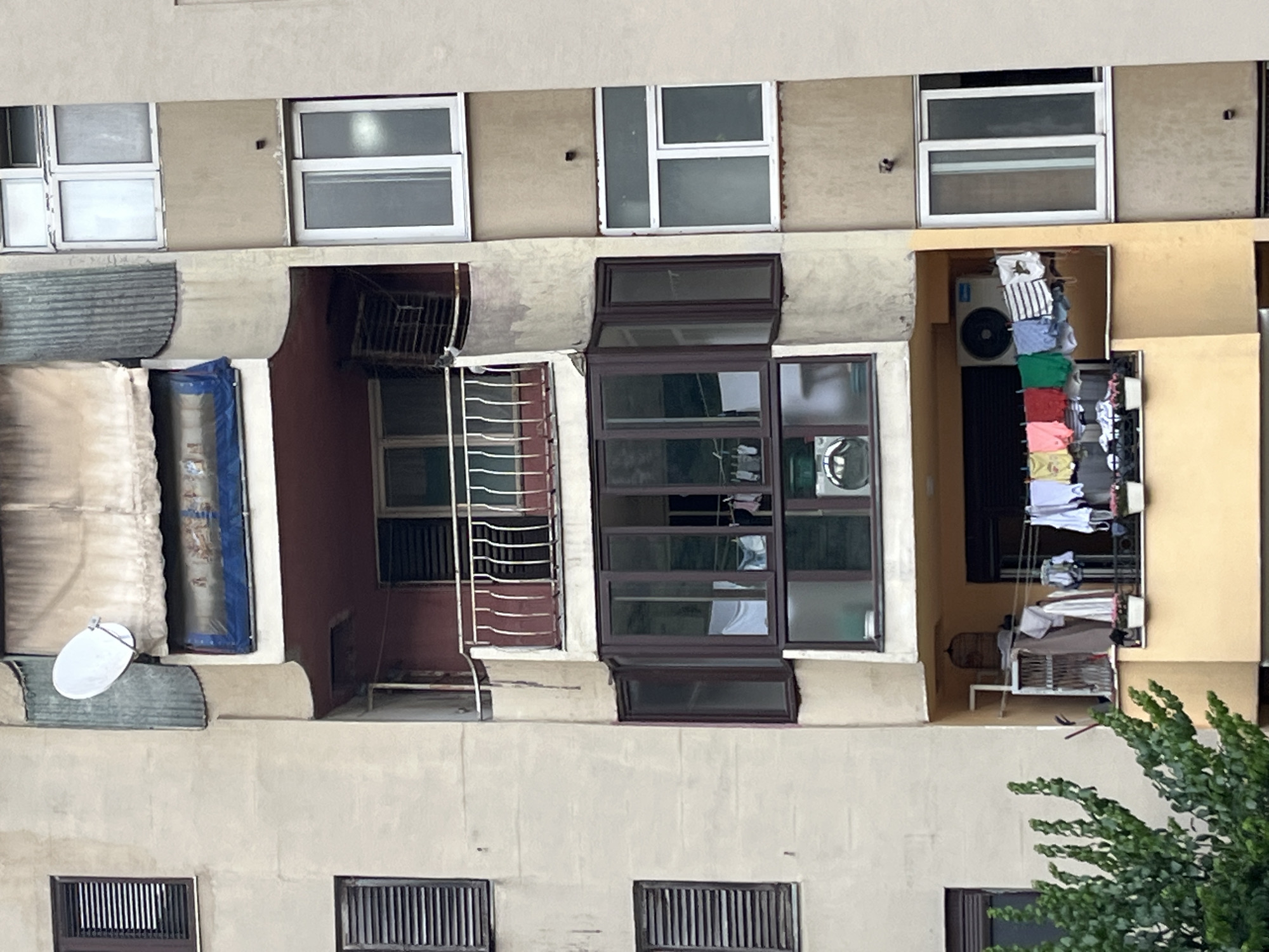 The image shows a close-up, eye-level, vertical shot of an apartment building facade in Škaljari, Montenegro. The building is constructed from light-colored concrete, with visible signs of wear and tear, including some staining and discoloration on the walls. The facade is segmented by balconies and windows, with the central section featuring a prominent bay window with dark brown framing, containing multiple panes. Inside this bay window, laundry is hanging on a line, including several items of clothing and a white sheet. Below this, on a lower balcony, more laundry is hung out to dry, featuring a variety of colored t-shirts, striped garments, and white items, alongside a clothes drying rack. An air conditioning unit is mounted on the wall above this lower balcony.
Several windows with dark brown shutters are visible on the left side of the building. On the right side, there are several windows with white frames. One of these, on the uppermost visible floor, has a satellite dish mounted on the exterior. The upper part of the building displays a section of weathered, off-white fabric awning supported by a corrugated metal structure. At the very top of the image, a white satellite dish is prominently positioned. Green foliage from a tree or bush encroaches on the lower left corner of the frame. The scene appears to depict a residential area, possibly in an urban or suburban setting, with everyday life activities like laundry drying being observed.