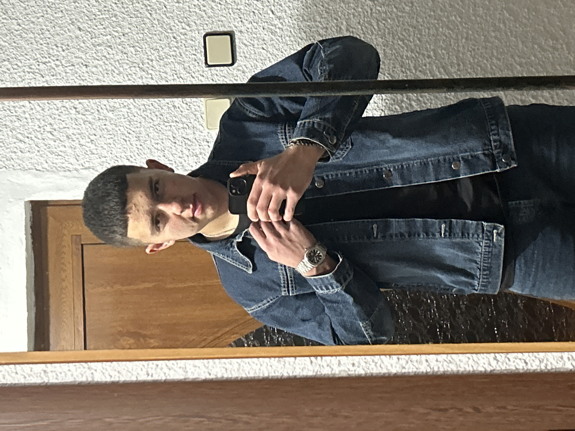 A young man stands in front of a mirror taking a selfie with his smartphone. He wears a denim jacket over a black shirt, denim pants, and a wristwatch. The reflection reveals a textured white wall, a light switch, a wooden doorframe, and a horizontal bar across the top of the mirror. The location is likely indoors, and the scene does not definitively indicate Zlatograd, Bulgaria.