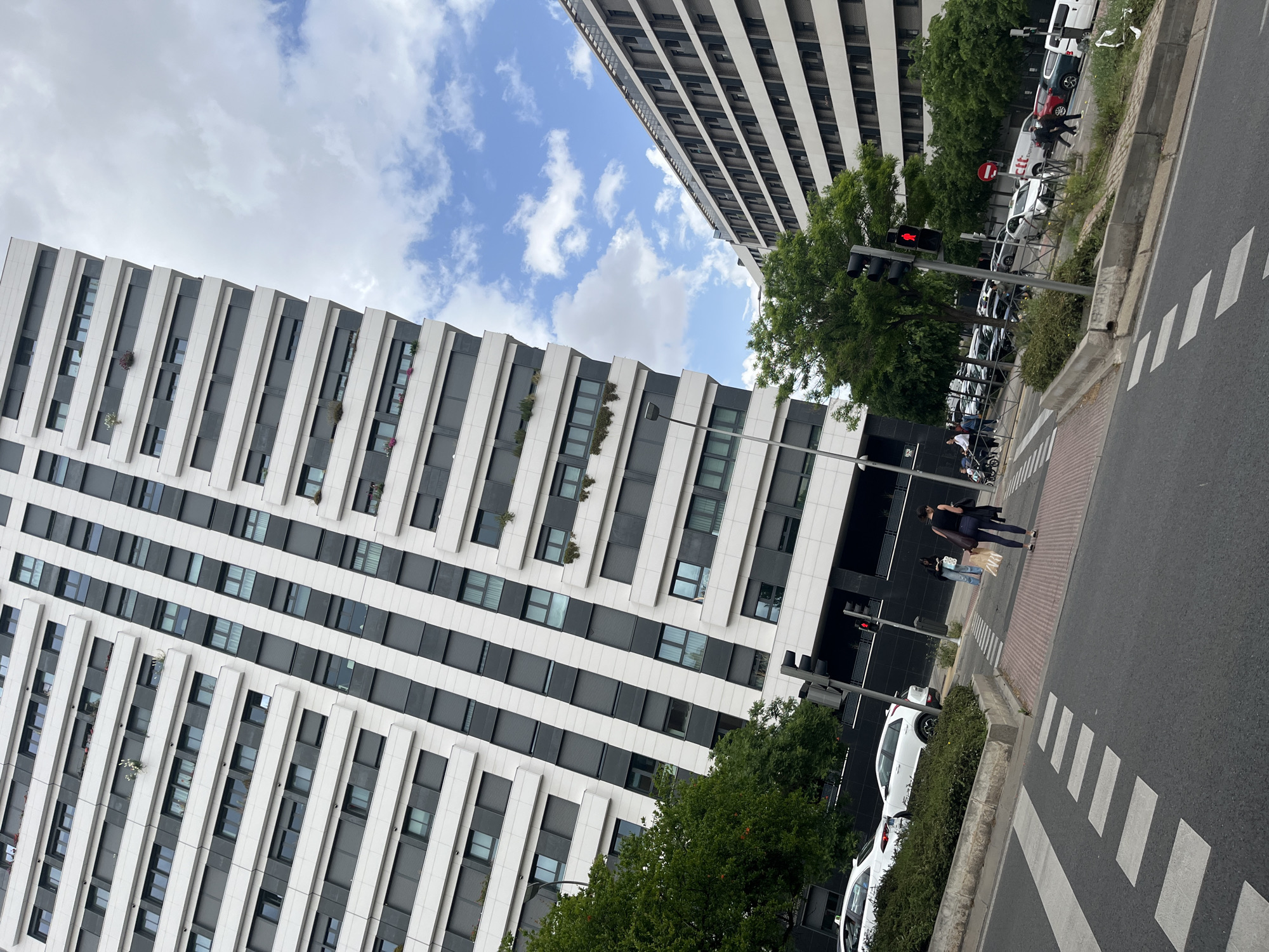 The image captures a modern urban street scene in Madrid, Spain, under a partly cloudy sky during daylight hours.
Two contemporary multi-story buildings frame the scene. The prominent building on the left features a striking facade of alternating vertical white panels and dark grey/black sections, likely windows or balconies. Several balconies on this building are adorned with small green plants. A second, similar building is partially visible in the upper right, distinguished by horizontal white and dark stripes.
In the foreground, an asphalt street with a clearly marked zebra crossing stretches across the lower right. A paved sidewalk runs parallel to the street, separated by a strip of green bushes. A row of parked vehicles lines the side of the street, including several white vans (one with "clt" written on its side), white cars, and a red car. Green trees are interspersed along the street and between the buildings. A set of traffic lights is visible, with the pedestrian signal illuminated red, indicating "do not cross."
Two women are seen walking on the paved sidewalk, approaching the zebra crossing. One woman, dressed in dark clothing, walks ahead, while the other, wearing light pants, walks behind her, carrying a white shopping bag with the "ZARA" logo clearly visible. Further down the street, another individual is on a scooter.
The scene depicts a typical moment of urban life in a modern city setting, characterized by its architecture, street infrastructure, and everyday activities.