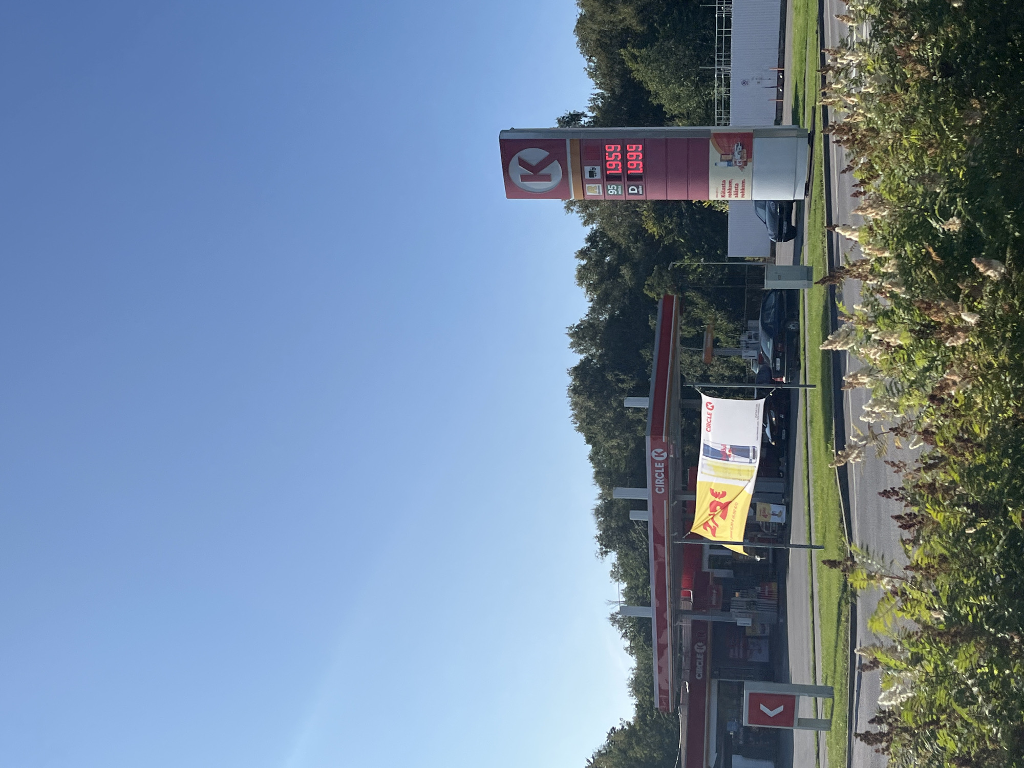 The image depicts a Circle K gas station on a clear, sunny day. A tall gas price sign shows prices of 1.959 for 95-octane gasoline and 1.999 for diesel (D). The gas station structure is red and white, with multiple pumps visible. A yellow and white banner displaying promotional information hangs in front of the station. Several cars are parked or driving at the station, and a white vehicle is visible in the distance. The foreground is filled with green foliage and flowering plants. The background consists of a dense line of trees. The location is identified as Kohtla-Järve linn, Estonia.