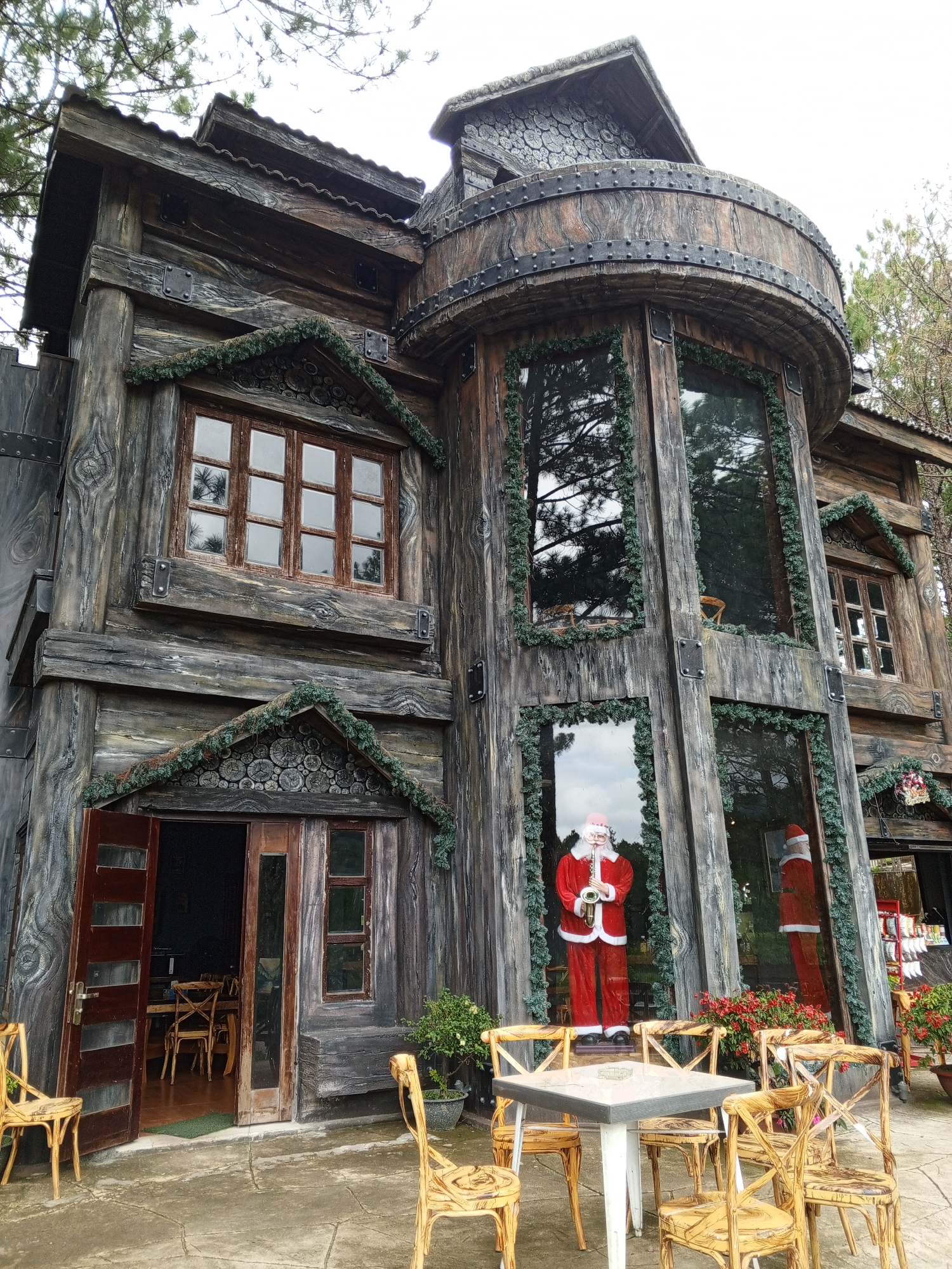 This is an exterior, eye-level, full-shot photograph of a unique, rustic-themed building in Da Lat, Vietnam, designed to resemble a large wooden log cabin. The building features a dark gray, wood-like texture with prominent vertical grain patterns and decorative metal bands. The facade includes multiple windows, some with multiple panes, and a prominent curved balcony on the upper level. Green garland is draped around the windows and along the roofline of a smaller gable above the entrance. The entrance is a double wooden door, one side ajar, revealing tables and chairs inside.
Outside the entrance, several light-colored wooden chairs are placed around a square white table. A potted plant with green foliage is situated near the entrance.
In front of a large window on the ground floor, a life-sized mannequin dressed as Santa Claus, holding a saxophone, is standing. Another Santa mannequin is visible in the reflection of a neighboring window. Red flowers are planted in front of the windows. The ground is paved with concrete. The architecture suggests a Christmas or winter theme.