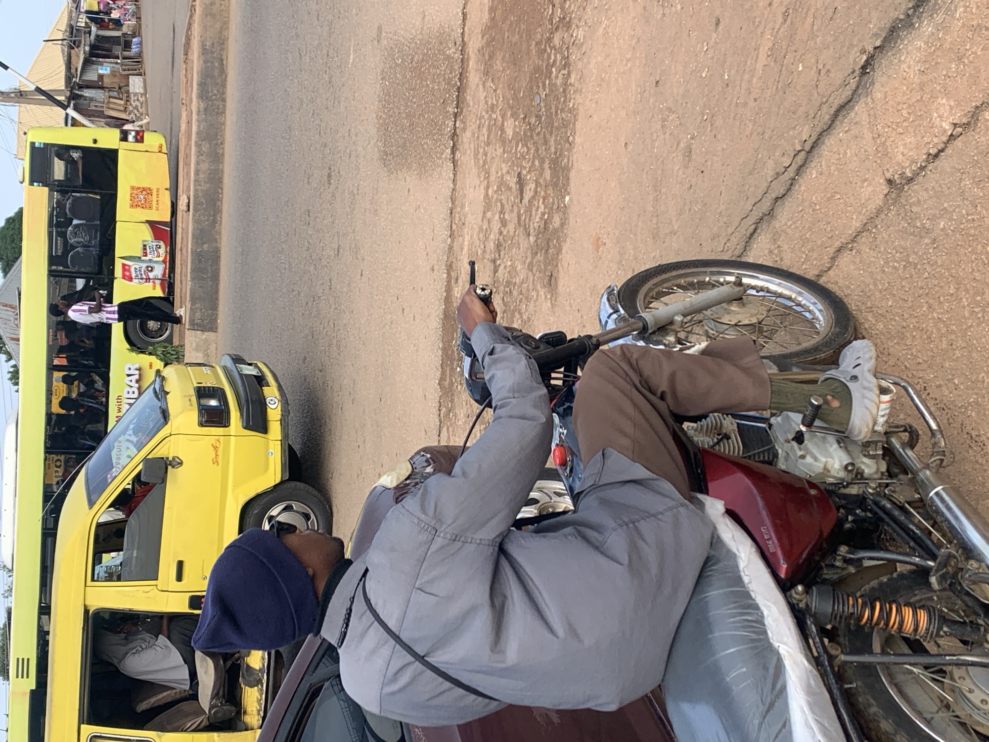 In Gura Top, Nigeria, a man wearing a blue beanie and a grey collared shirt rides a red and black motorcycle. He is wearing brown pants and grey crocs. The motorcycle has a black seat covered in plastic. The man is looking to his left. Behind him, a yellow van and a larger yellow bus are parked on the side of the road. The bus has advertisements on its side. The road appears to be paved and is cracked in places. The environment suggests a street scene in a town or city.