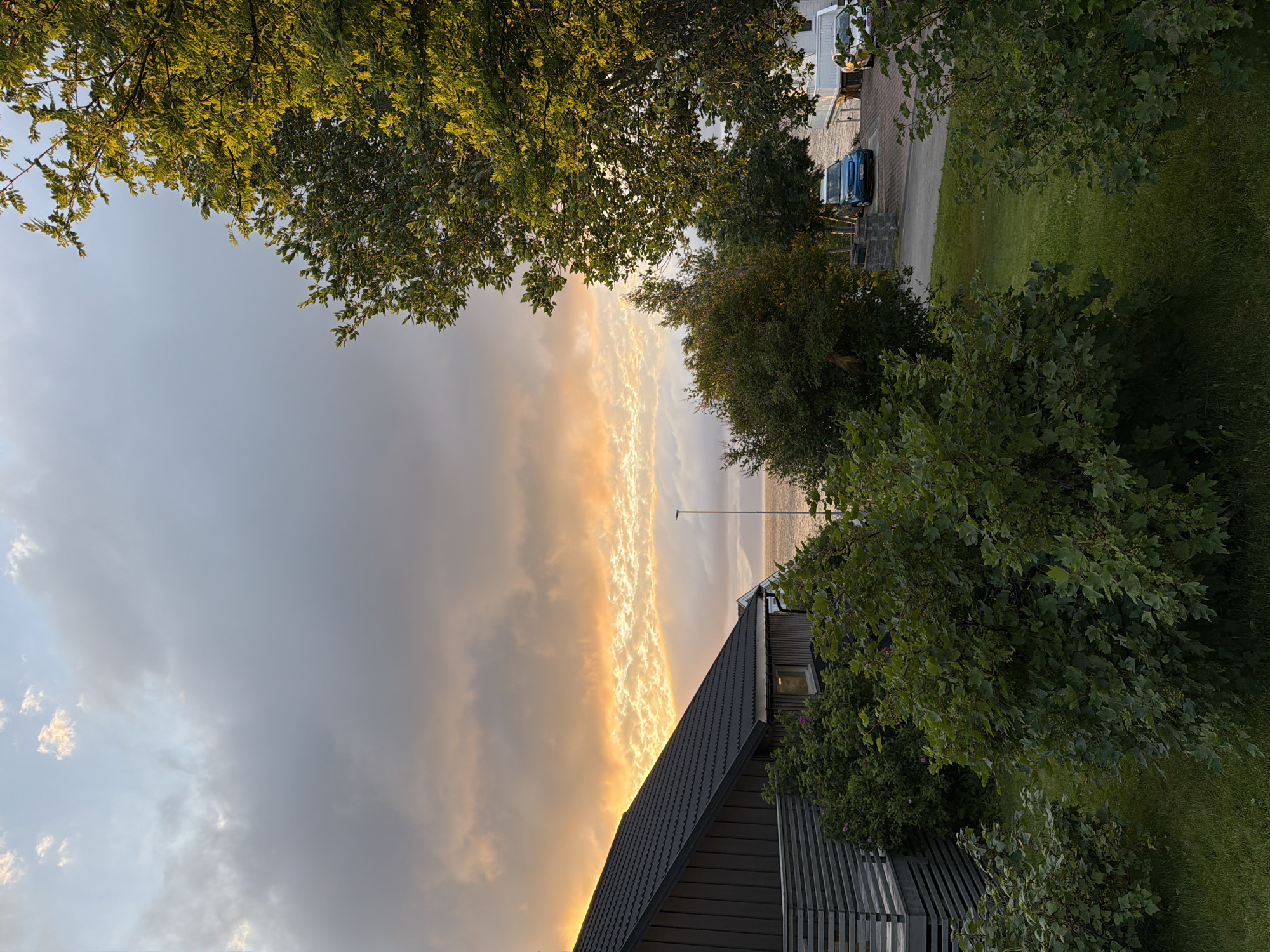 A photograph captures a coastal scene at dusk, likely in Harstad, Norway, given the architectural style and natural landscape. The sky is dominated by dramatic clouds, with a band of bright orange and yellow light illuminating the lower portion, suggesting a sunset. To the left, a modern dark-colored house with a sloped roof and a grey wooden fence is visible. Lush green bushes and trees are scattered throughout the foreground and mid-ground. In the mid-ground, a street runs parallel to the water, where a blue electric car is parked next to a white house. The water, appearing calm, stretches to the horizon, with a distant landmass or mountain silhouetted against the bright sky. The overall lighting is soft and warm, characteristic of the golden hour. There are no people or animals visible in the frame.