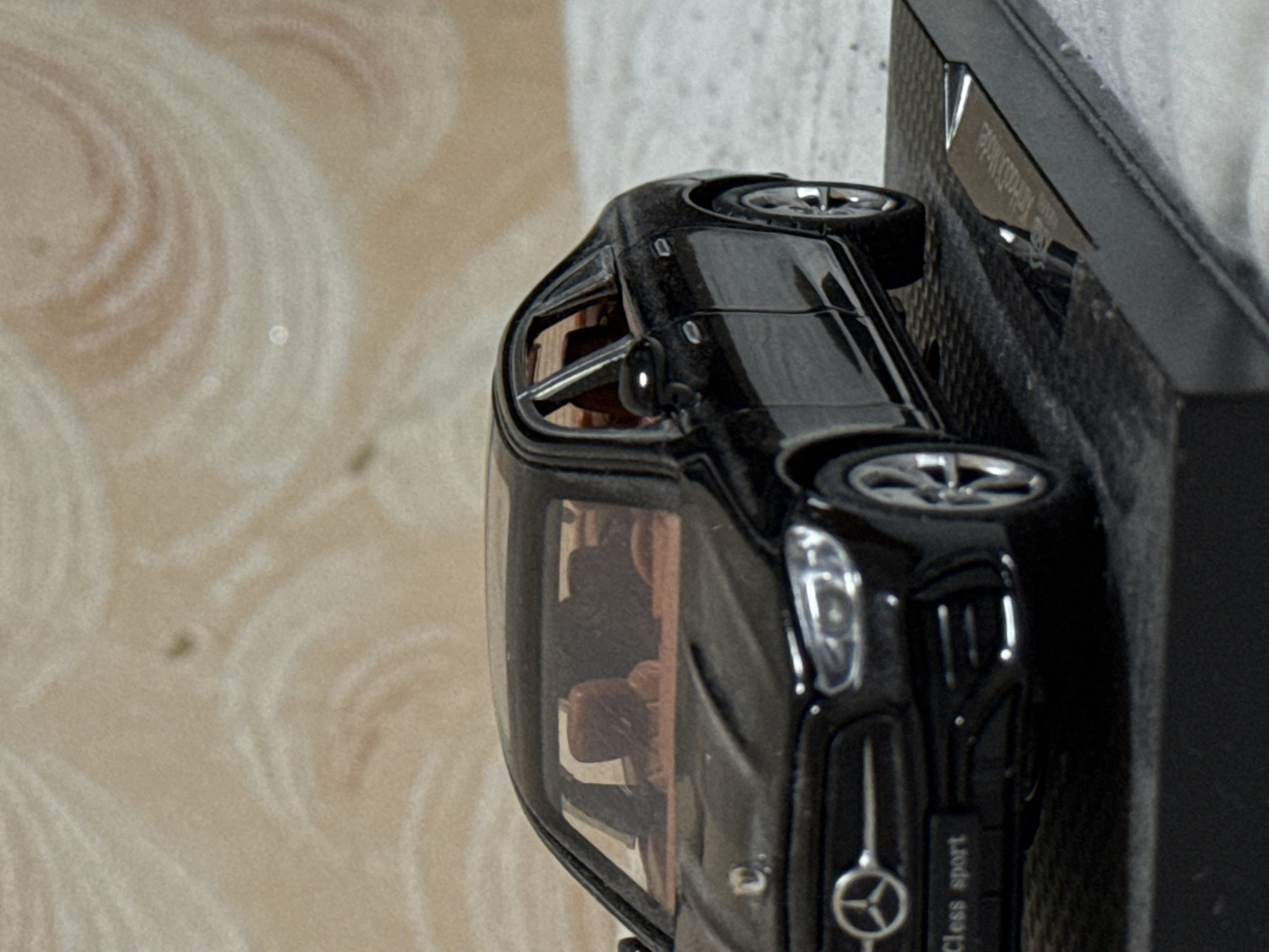 Here is a detailed description of the image:
The image shows a black Mercedes-Benz C-Class Sport model car displayed on a black, possibly carbon fiber textured, stand. The model car is positioned diagonally, with its front end facing the lower right corner of the image. The stand appears to be on a surface with a light tan, swirled pattern, potentially a tabletop or display surface. The car's details, such as the wheels, headlights, and the Mercedes-Benz logo on the front, are visible. The interior appears to have a light brown color. The location in the city of Лабжанскі сельскі Савет, Belarus is not verifiable from the image.