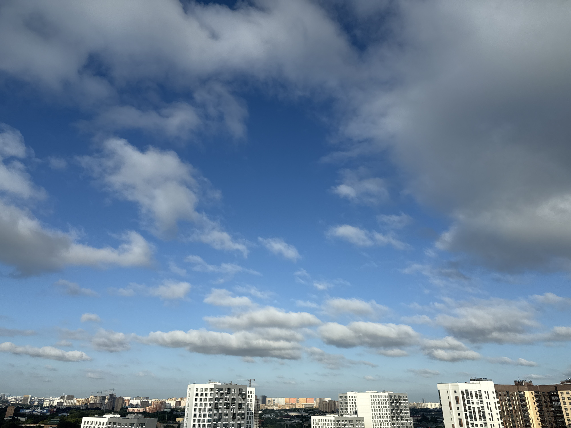 A city skyline in Lyubertsy, Russia, is visible under a dynamic sky with scattered clouds. Several multi-story residential buildings are prominent in the foreground, characterized by modern architecture with white and dark facades, many featuring a grid of windows. In the mid-ground, a dense urban landscape unfolds with a mix of buildings of varying heights and colors, including some under construction with visible cranes. The background shows a hazy horizon, suggesting the vastness of the city. The sky is a vibrant blue, partially obscured by large, wispy white clouds and larger, darker, storm-like formations on the right side of the frame. The scene appears to be captured from a high vantage point, overlooking the urban sprawl. There are no visible people or vehicles in the image.