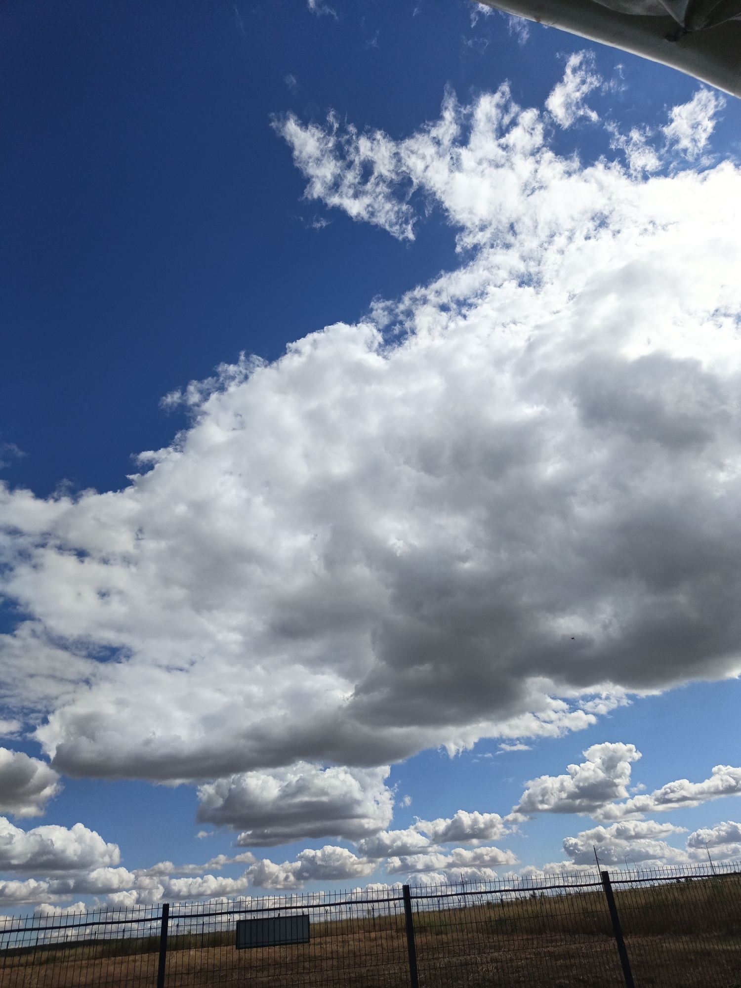 A wide-angle, slightly upward-facing shot captures a vast expanse of blue sky filled with numerous clouds. The clouds vary in size and density, ranging from wispy formations to large, towering cumulus clouds. A portion of a white structure, possibly a canopy or awning, is visible in the upper right corner, casting a shadow that is not depicted in the frame. In the lower third of the image, a dark, wire-mesh fence runs horizontally across the frame. Beyond the fence, dry, brown grass and some sparse vegetation are visible. A dark rectangular sign or panel is affixed to the fence in the middle ground, slightly to the left of center. The scene appears to be an outdoor location, possibly a field or open area, with a fence marking a boundary. The lighting suggests it is daytime, with sunlight illuminating the clouds from above and to the side, creating bright white highlights and darker shadowed areas within the cloud formations. There are no people or animals visible in the frame. The location is described as Unknown, Unknown.