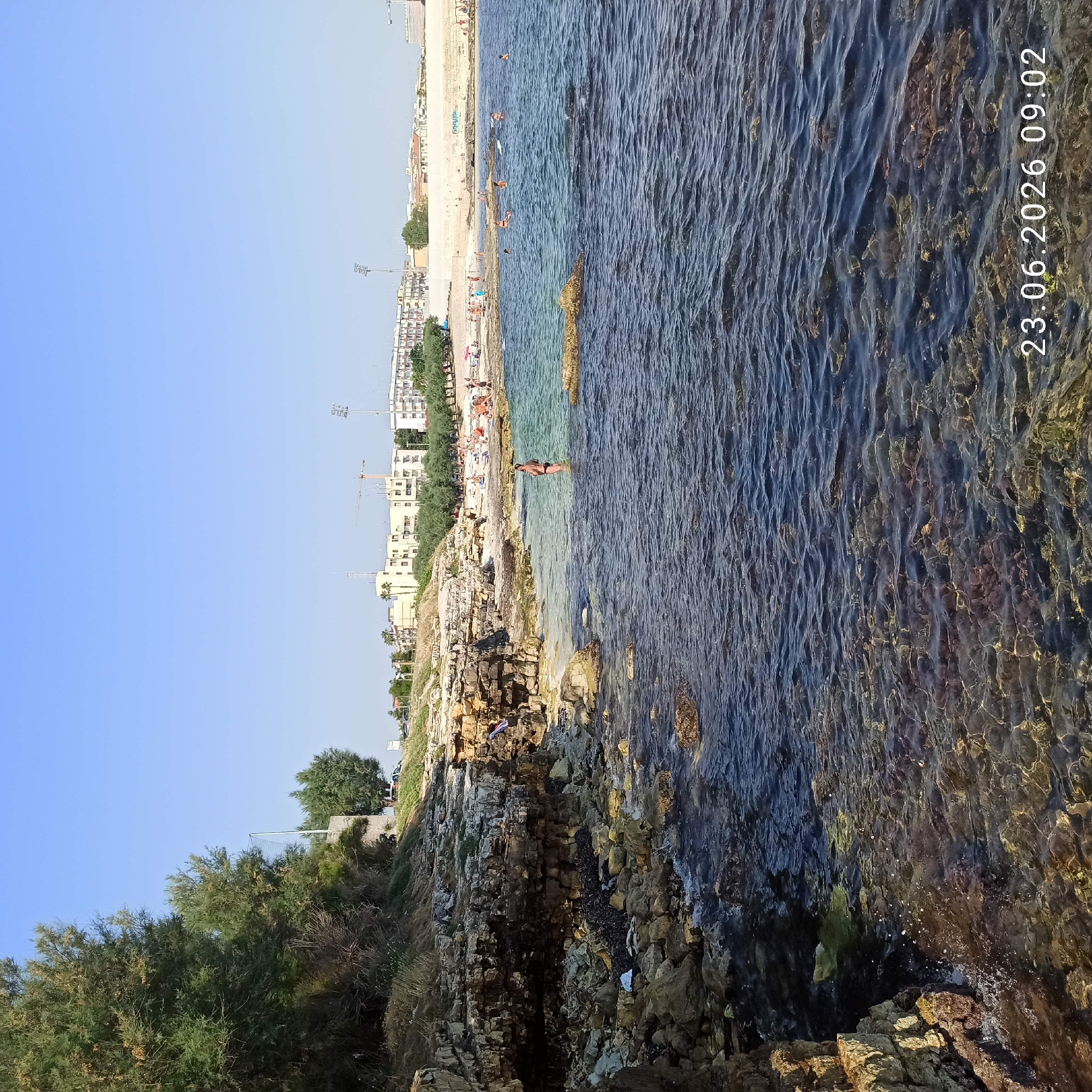 A coastal scene in Molfetta, Italy, features a rocky shoreline with a clear blue sky overhead. The water is a vibrant turquoise near the shore, gradually deepening to a darker blue. In the water, several people are visible, some wading and others swimming. A woman in a black bikini stands in the shallows. Further out, a group of people are sunbathing on a sandy beach, backed by trees and buildings that suggest an urban or resort area. The rocky coastline is covered in patches of vegetation and seaweed. The image is dated "23.06.2026 09:02."