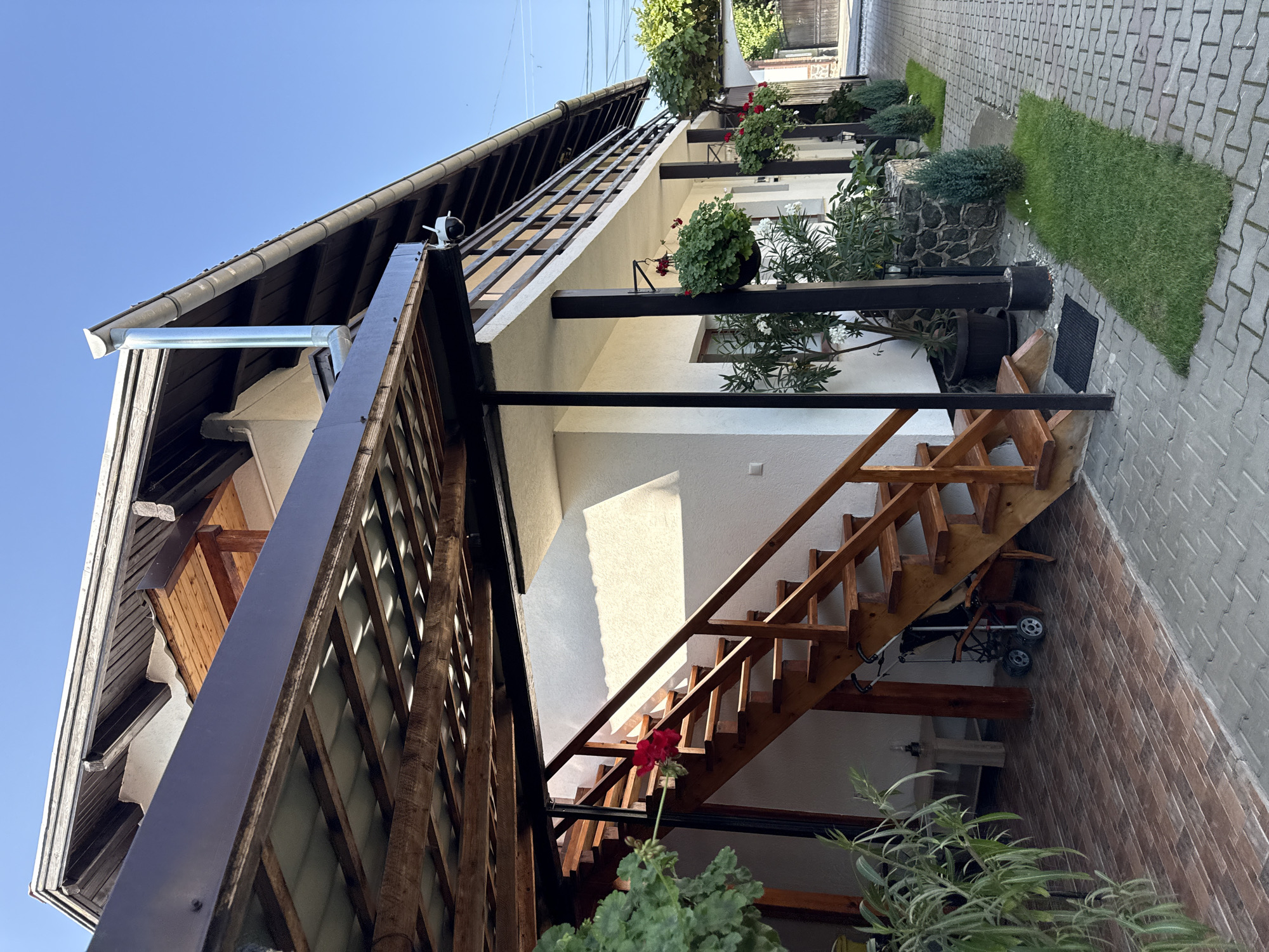 The image depicts an outdoor scene at a residence in Cârțișoara, Romania. The foreground shows a paved walkway with interlocking grey pavers, transitioning to a tiled area near a wooden staircase. The staircase, made of light-colored wood, leads up to a balcony. Green plants, including potted shrubs and a vine, are visible throughout the scene.

The building has white stucco walls and a dark-colored, sloped roof with visible wooden beams. A metal downspout extends vertically from the roof. A security camera is mounted on the exterior of the building. Hanging baskets with red flowers are suspended from the balcony railing and near the base of the building.

In the middle ground, a baby stroller is parked at the base of the stairs. In the background, the walkway continues towards a gateway, with more greenery and a glimpse of a fence. The sky is clear and blue, indicating fair weather. The overall impression is of a well-maintained residential property.