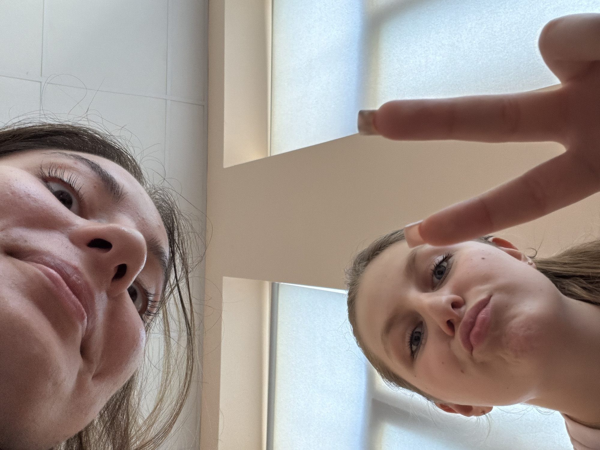 In a close-up, low-angle selfie, two young women are featured. The woman in the foreground, on the lower left, looks directly at the camera with her mouth pursed and raises two fingers in a peace sign. Her hair is pulled back into a ponytail, and she is wearing a light pink shirt. To the right of her face, her hand is prominently displayed, with painted nails on her fingers. In the upper portion of the image, the face of another young woman is visible, upside down and cropped. She has dark hair that falls around her face, and her eyes are open. The background consists of a white ceiling with grid lines and a window with sheer curtains. The scene appears to be indoors, possibly a school or home environment. There are no discernible elements to indicate the location within Burgas, Bulgaria.